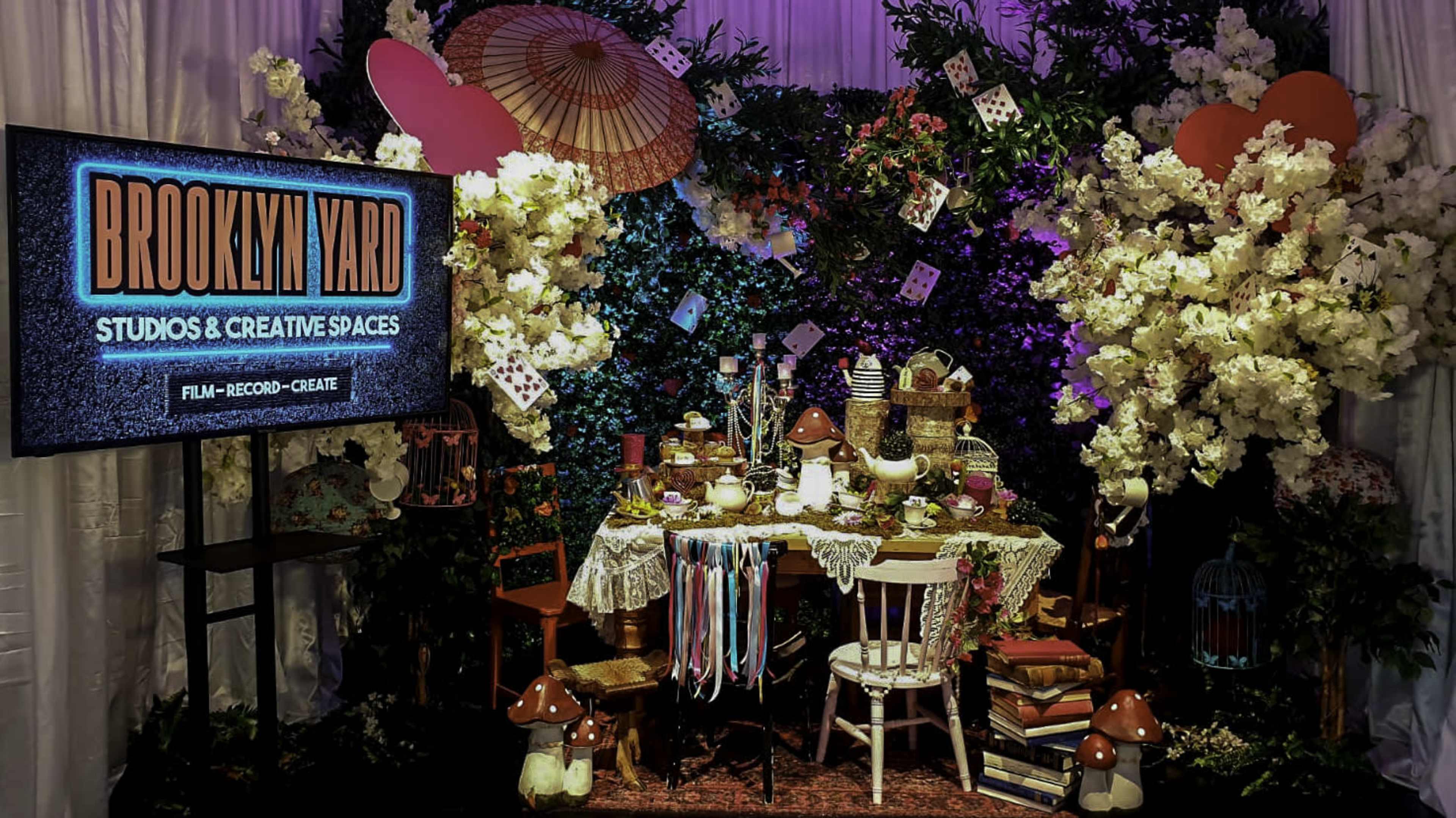A decorated studio space features a table set with various items surrounded by lush greenery and floral arrangements, with a sign that reads "BROOKLYN YARD STUDIOS & CREATIVE SPACES."