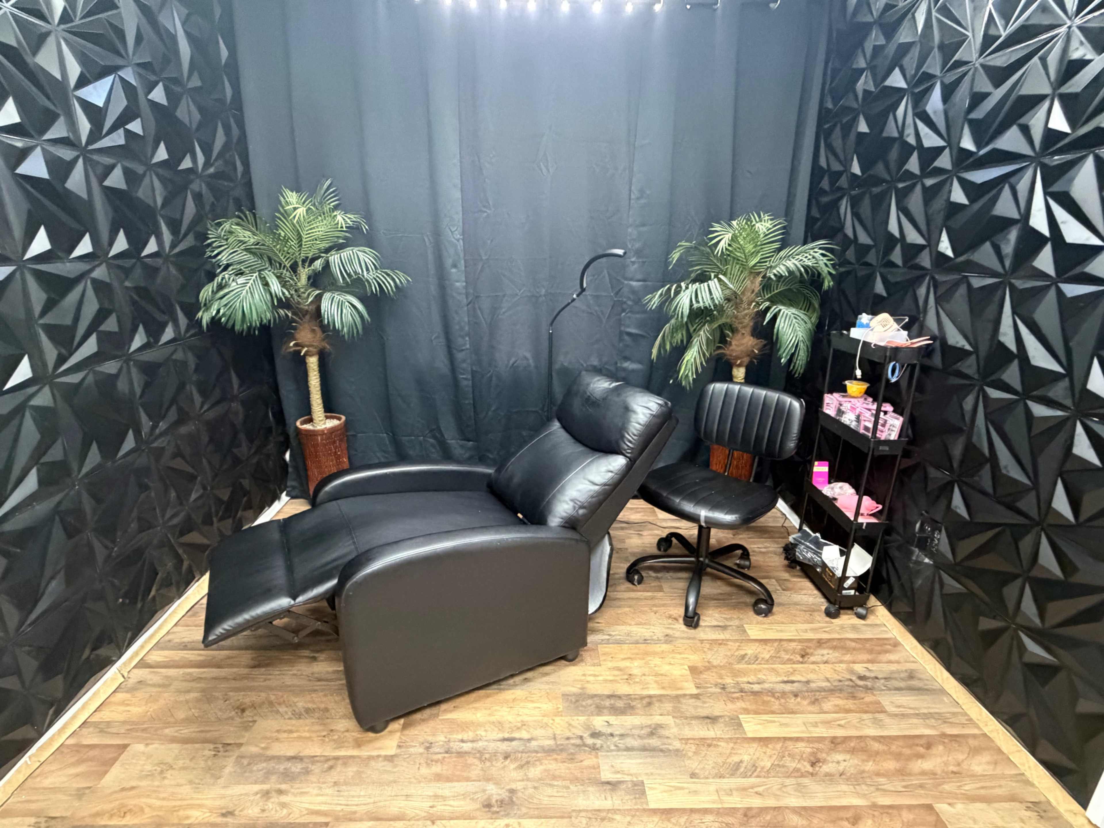 The image shows a small room with a black reclining chair, a black rolling office chair, two potted faux palm trees, and a shelf containing various items against a textured black wall.