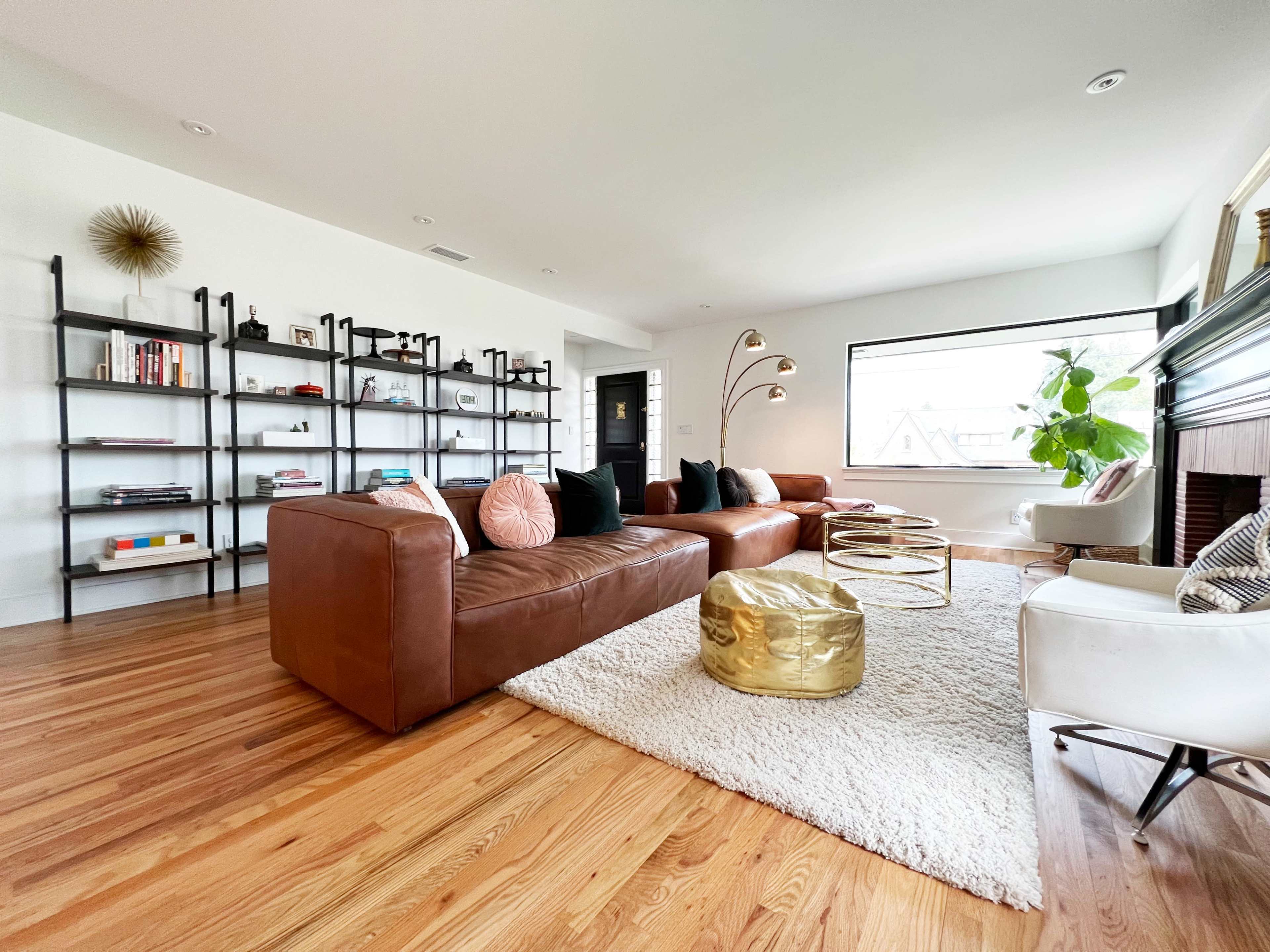 The living room features brown leather sofas, a white rug, a gold accent table, and a large window illuminating a modern bookshelf.