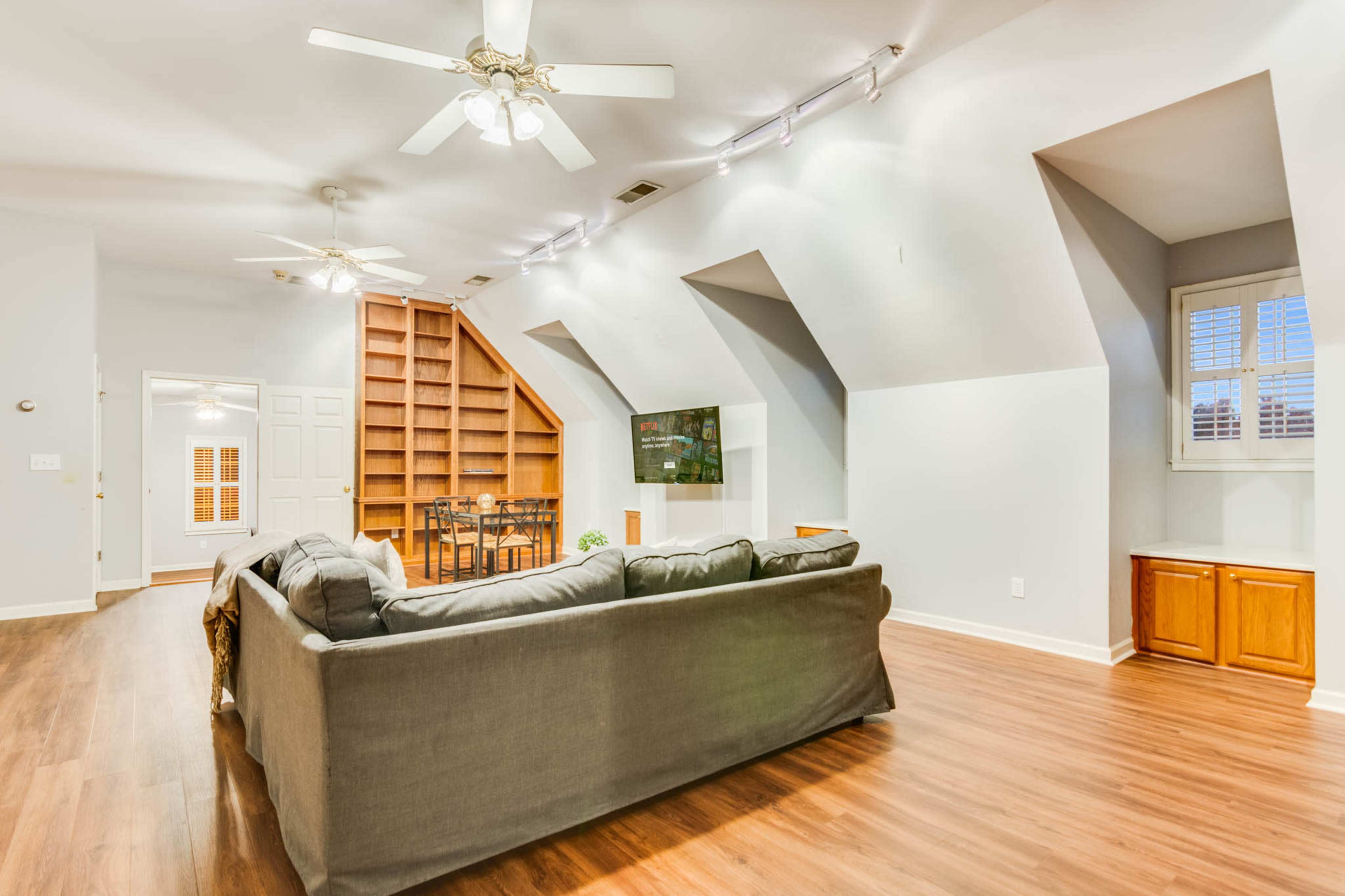 The image shows a spacious living room featuring a gray couch, a mounted TV, and a dining area with a wooden table, all under sloped ceilings with ceiling fans.