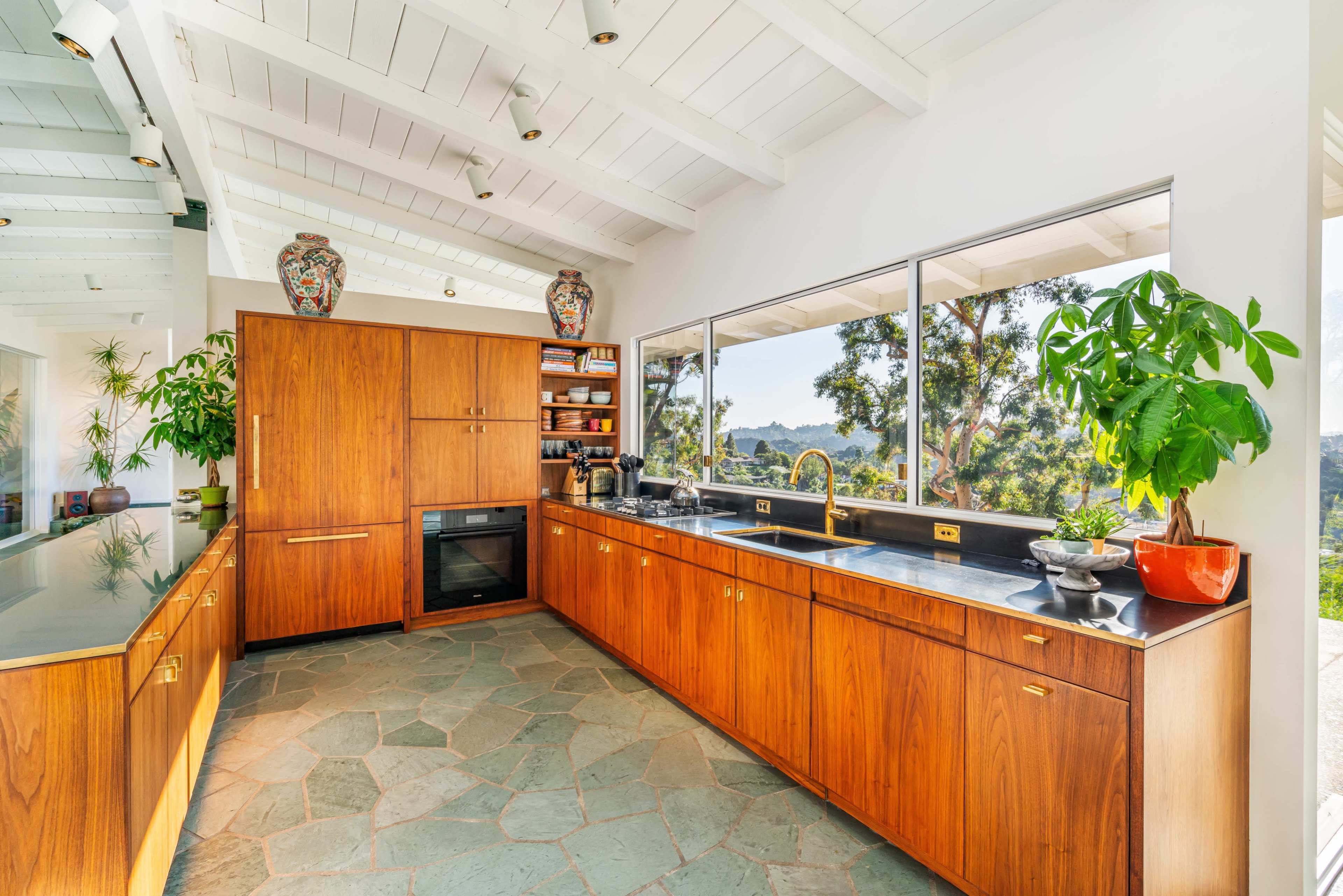 A modern kitchen features wooden cabinetry, a stone floor, and large windows showcasing a view of the outdoors.