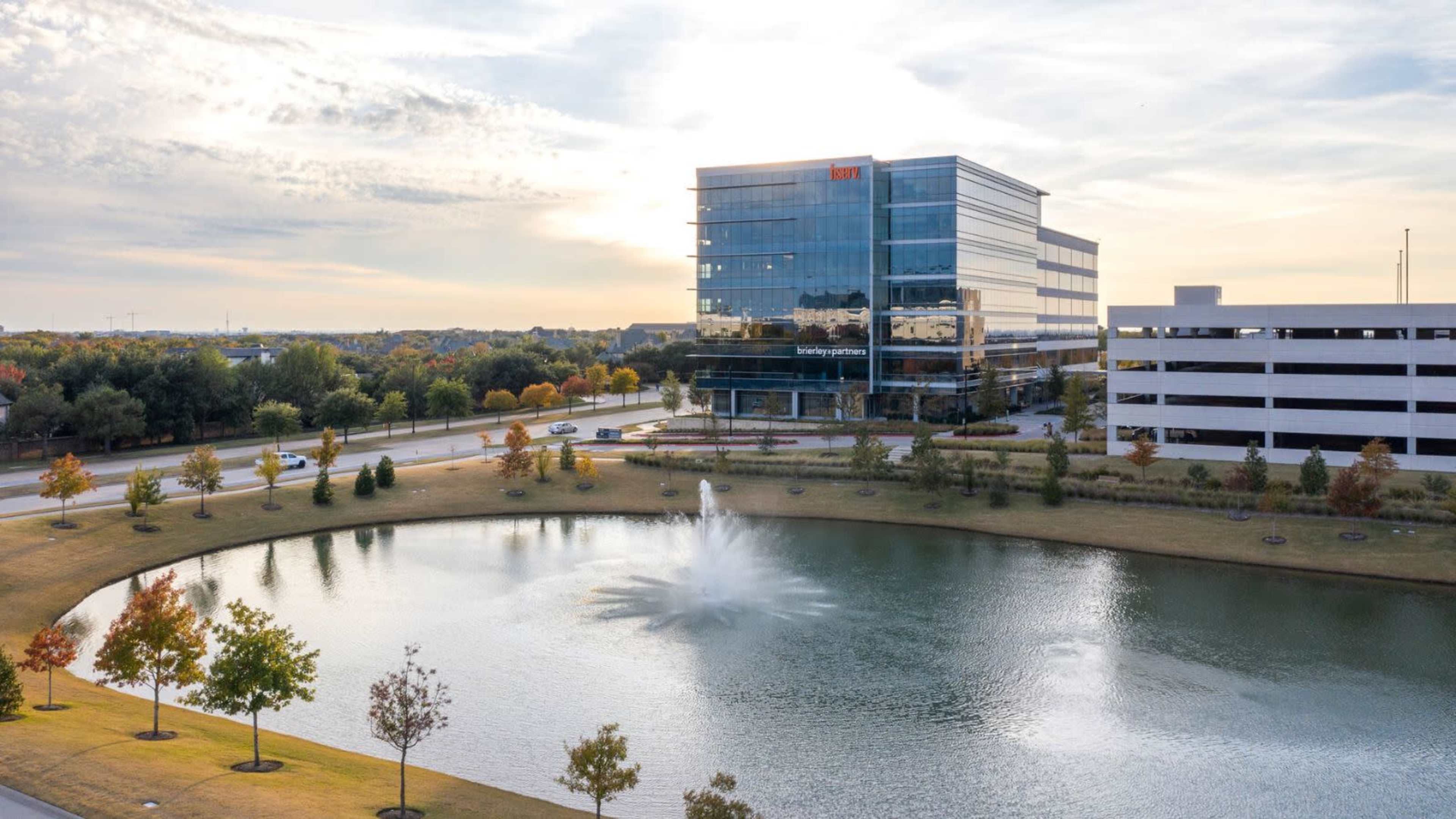 A modern office building is situated next to a pond with a fountain, surrounded by landscaped greenery and roads.
