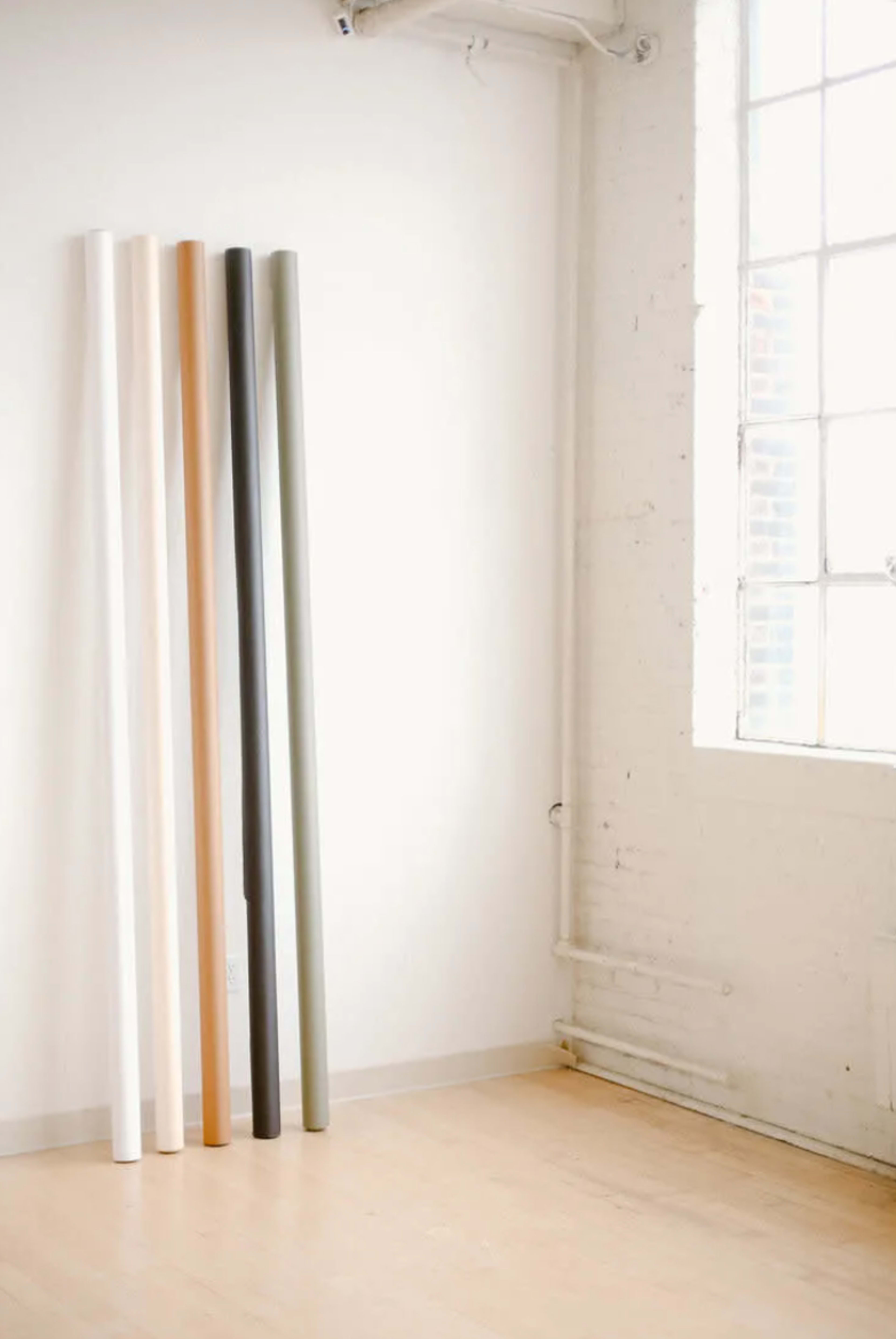 Five rolls of paper in various colors are standing upright against a white wall beside a large window.