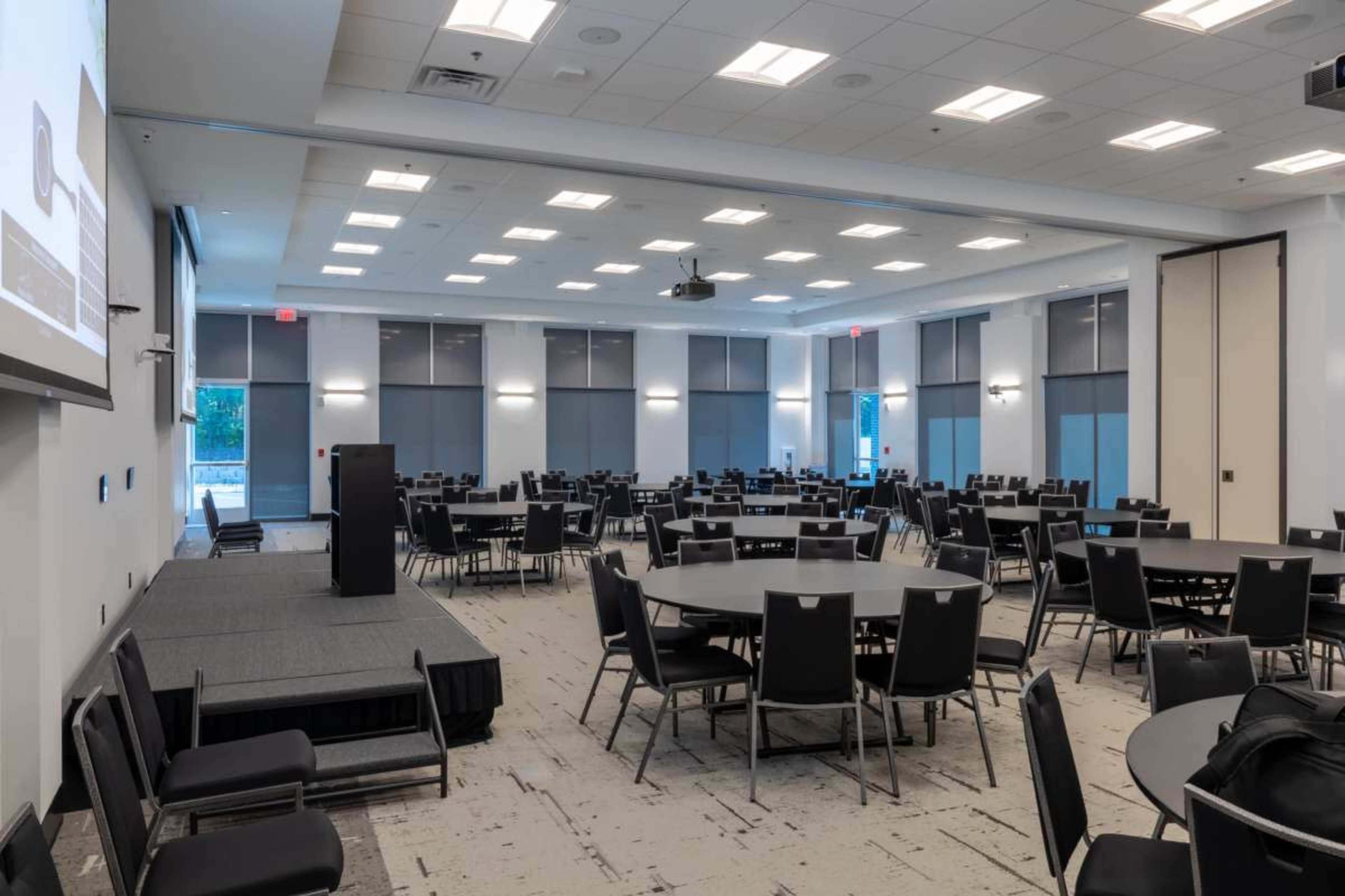 A spacious conference room features multiple circular tables arranged around a stage with a podium, illuminated by natural light from large windows.