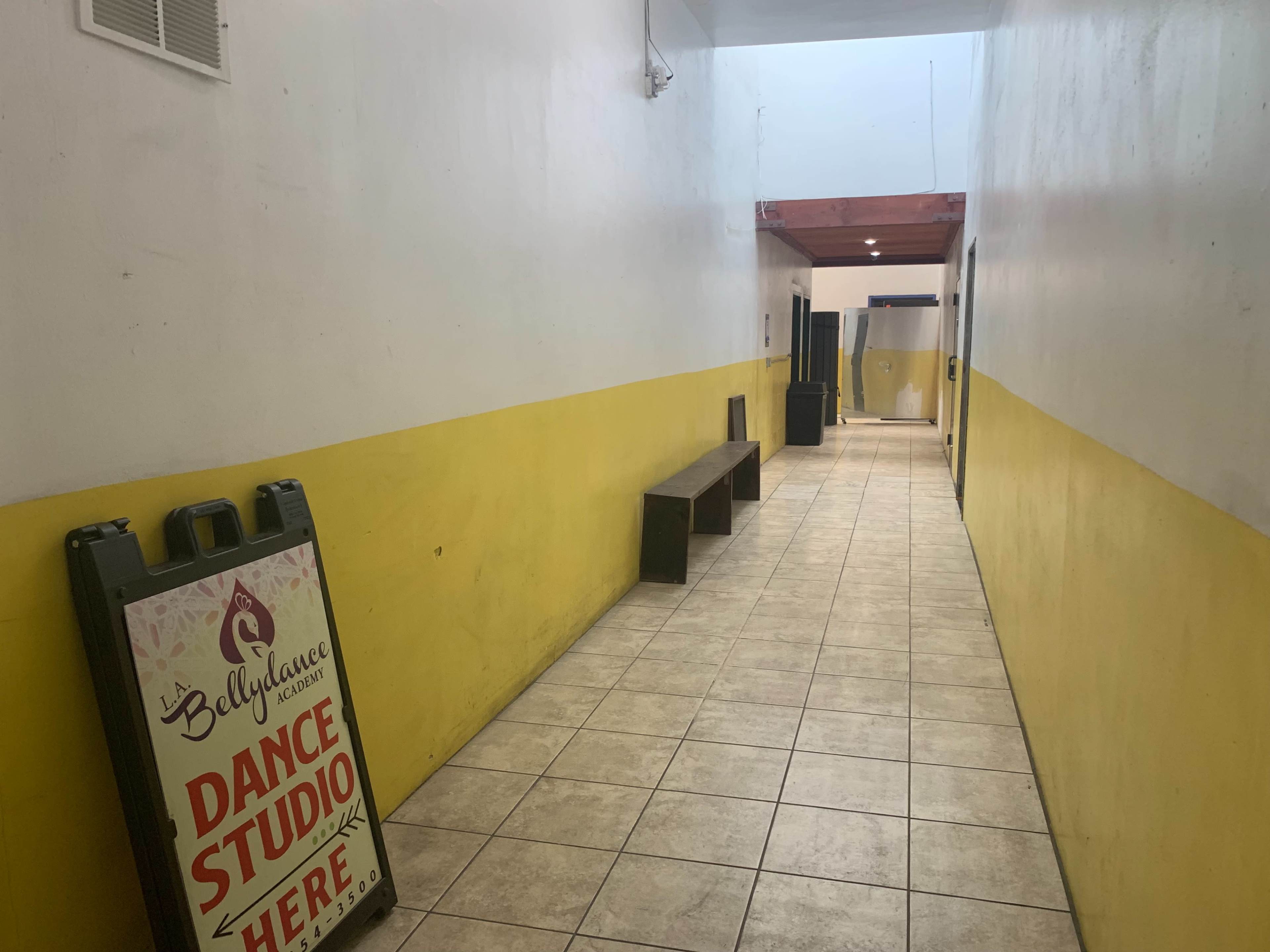 The image shows a long hallway with yellow and white walls, featuring a dance studio sign on an easel, benches along one side, and doors leading to other rooms.