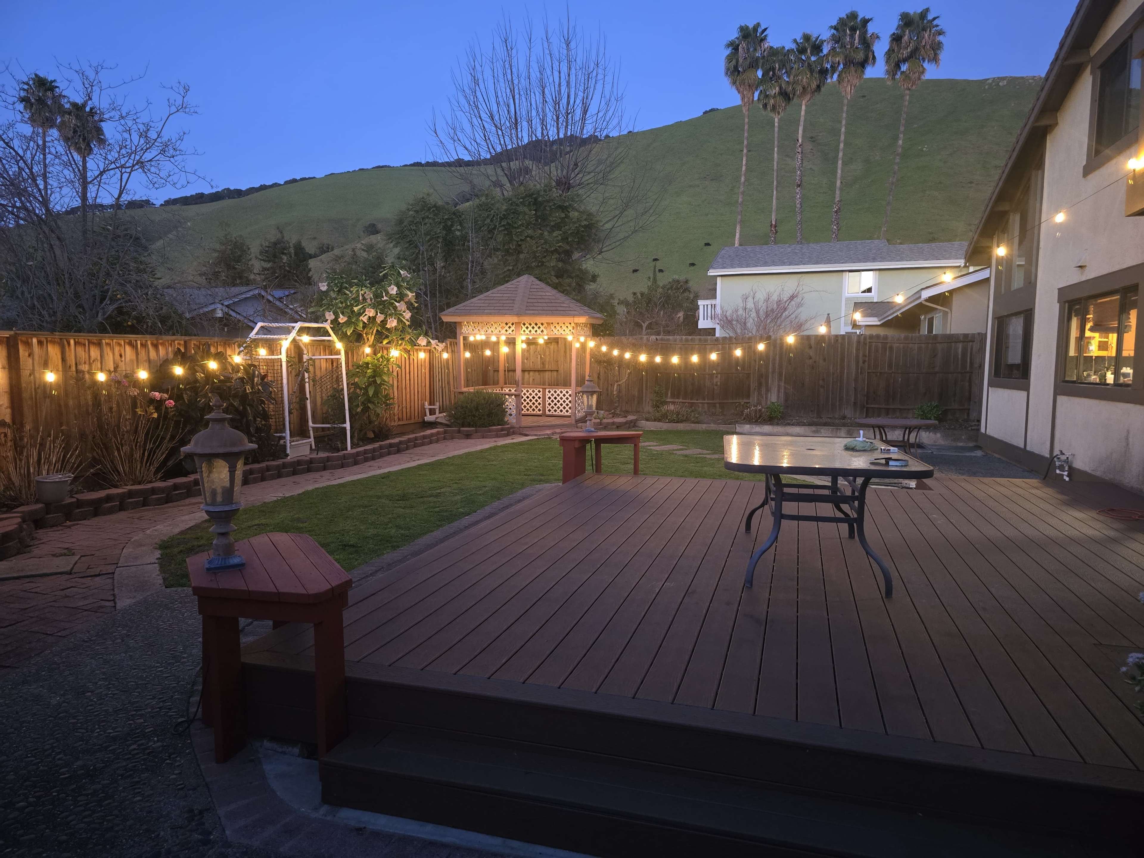 The image shows a backyard patio with wooden decking, a gazebo, and string lights, set against a backdrop of a green hillside at dusk.
