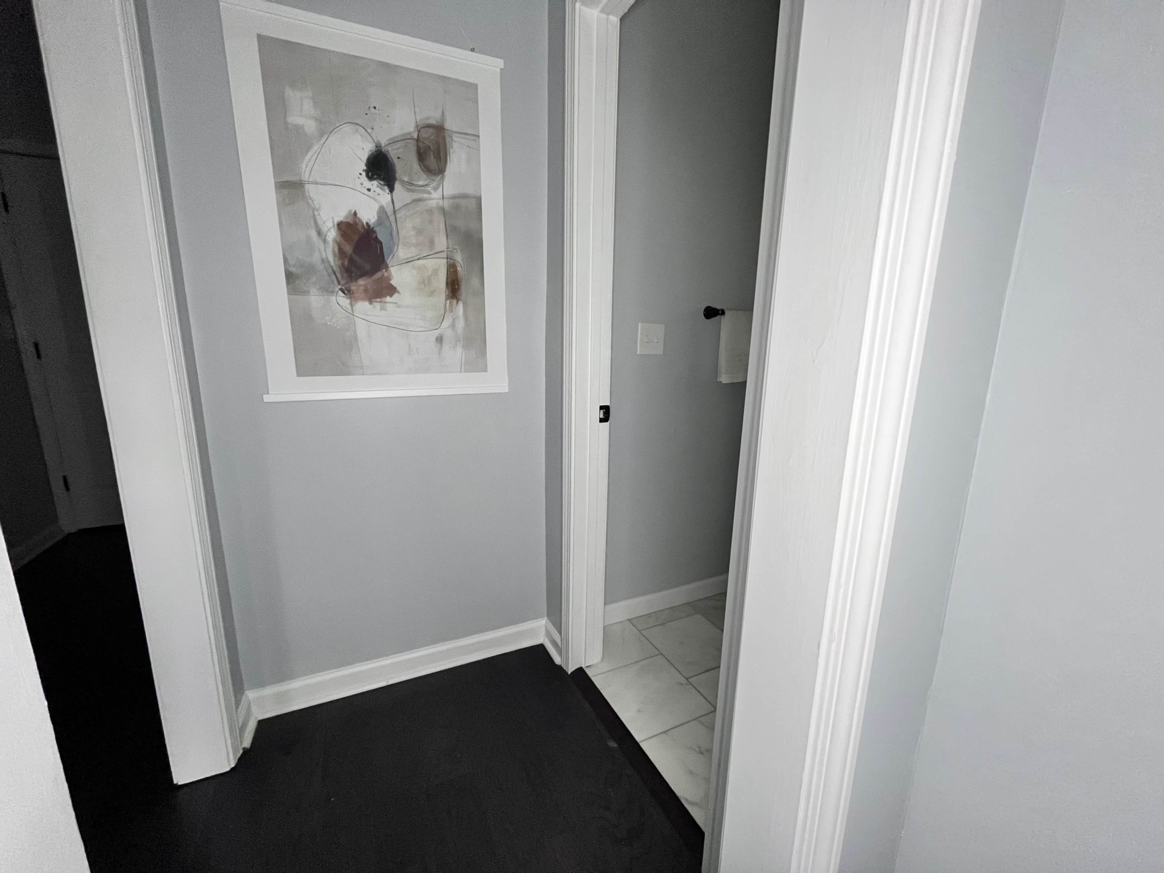 The image shows a narrow hallway with gray walls, a framed abstract artwork on one wall, and a bathroom door partially open at the end.