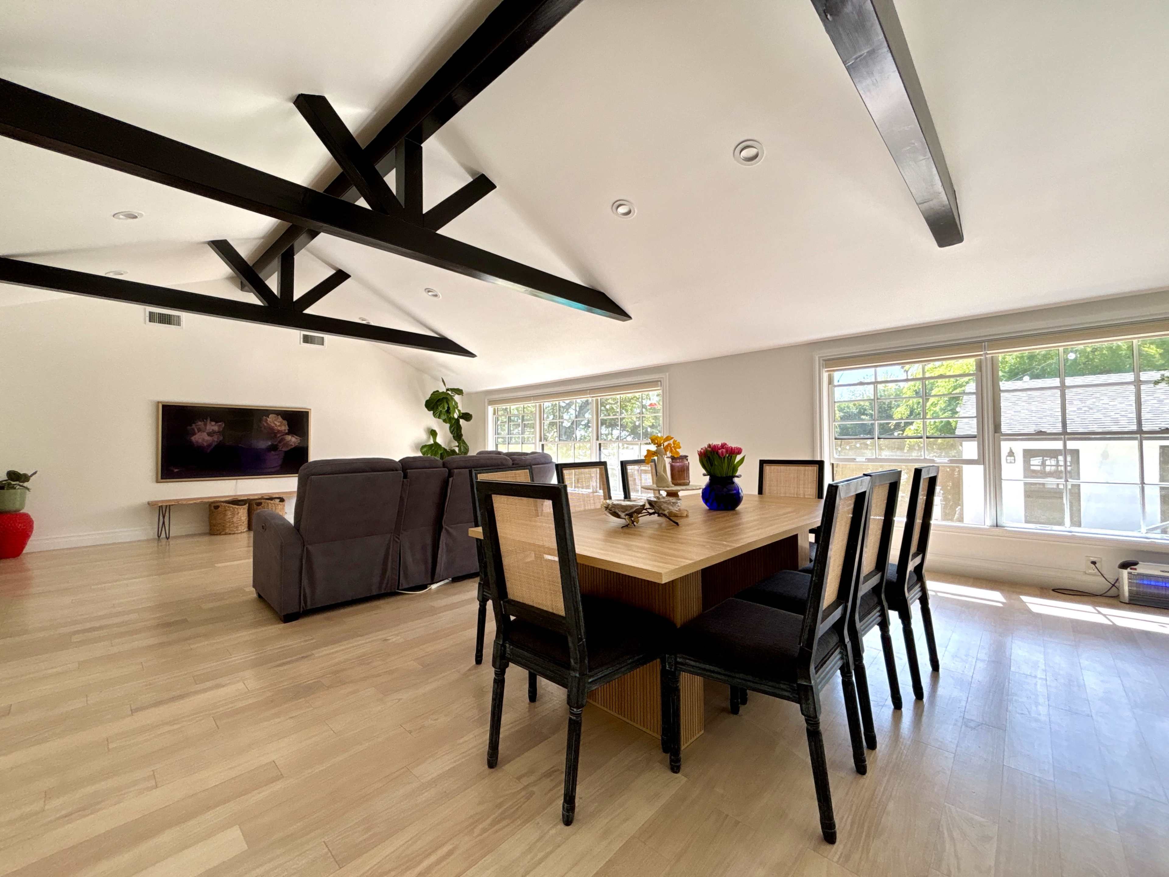 The image shows a modern living space featuring a dining area with a wooden table and black chairs, adjacent to a seating area with gray sofas and a television mounted on the wall.