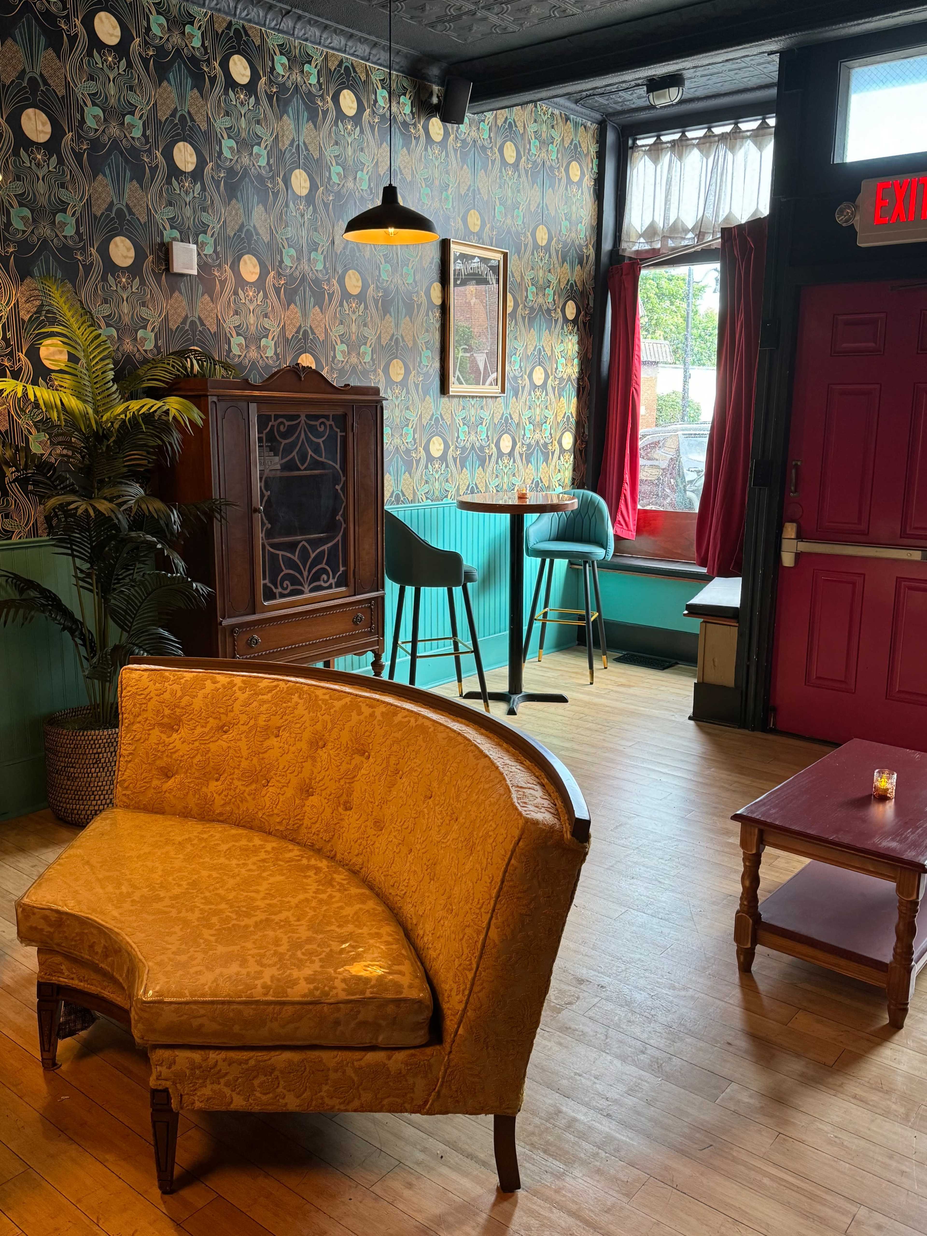 A cozy lounge area with a yellow curved sofa, a small wooden table, and a bar counter in the background next to a window.