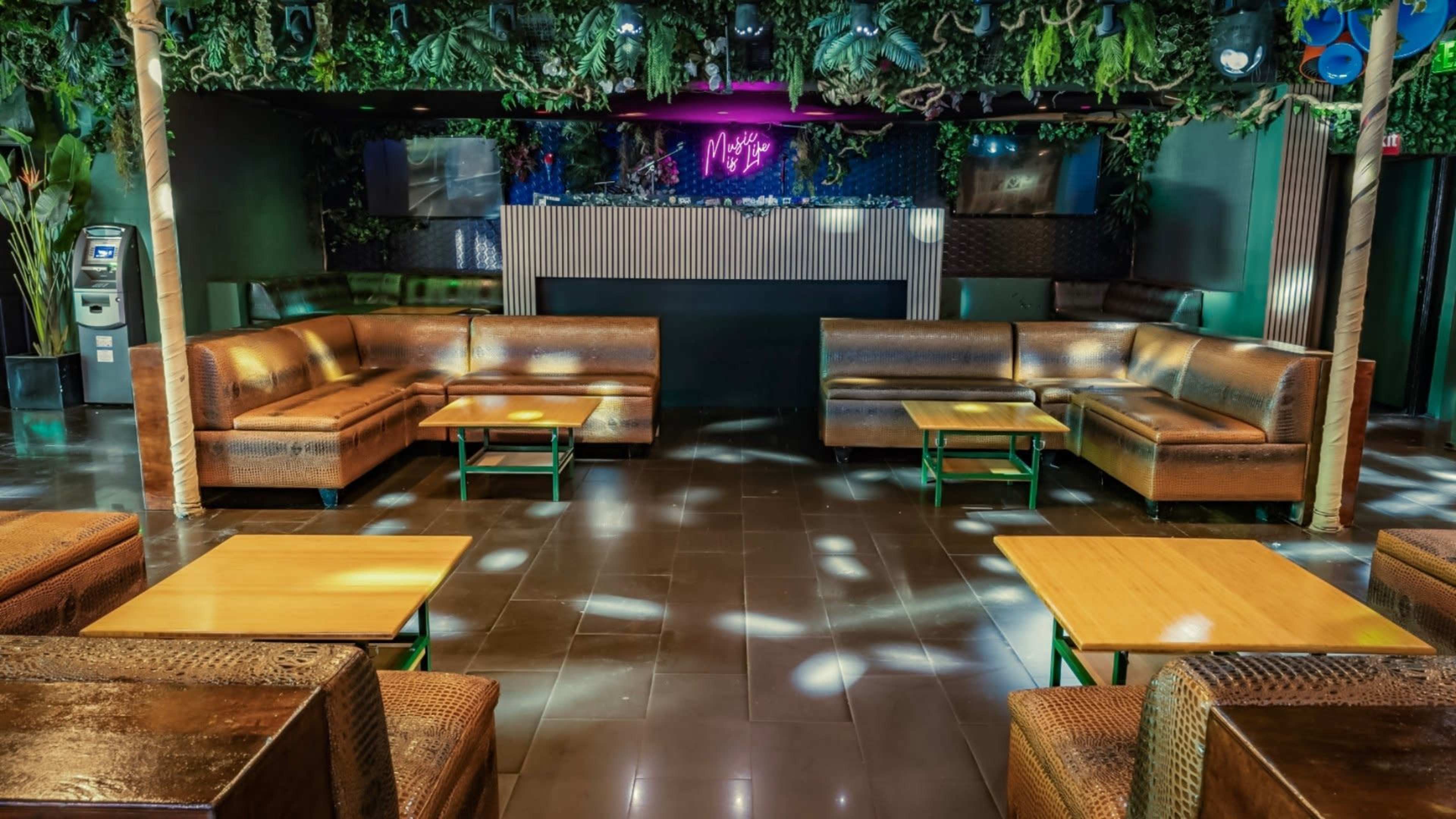 The image shows a modern lounge area with brown leather seating, wooden tables, and greenery overhead, set under dim lighting.