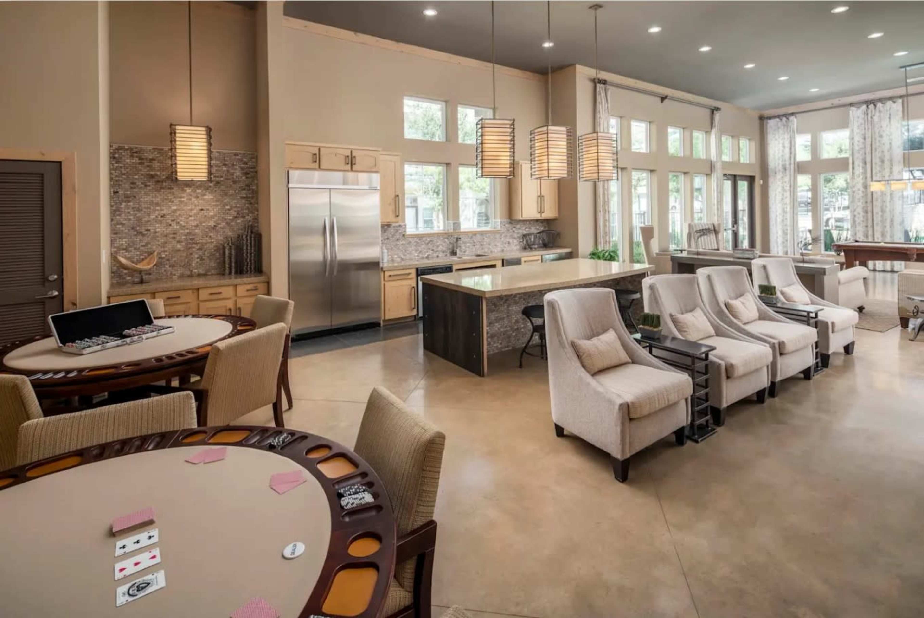 The image shows a spacious, modern interior of a room featuring a card table, a kitchen area, and comfortable seating arrangements with large windows allowing for natural light.