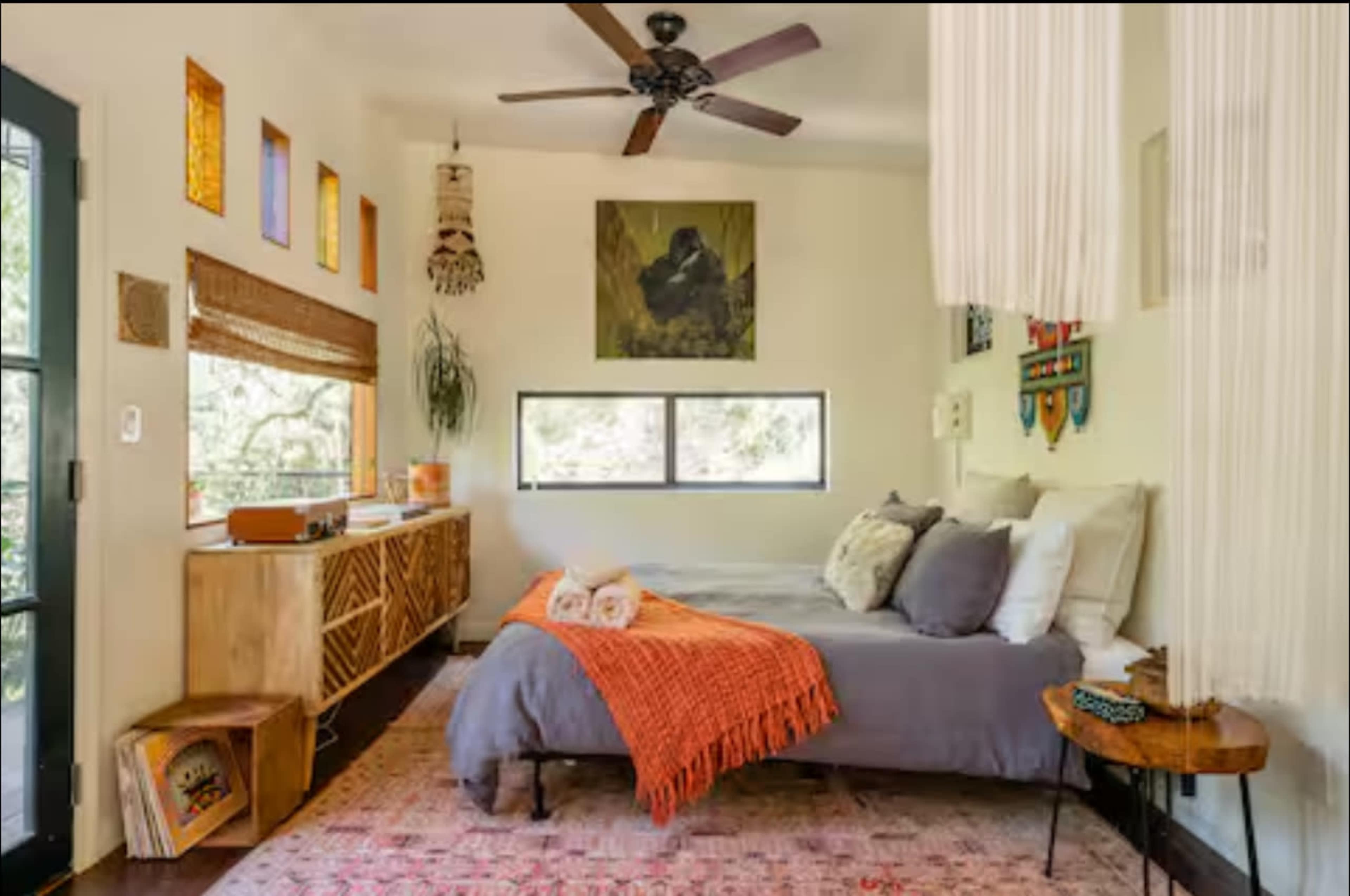 The image shows a cozy bedroom featuring a bed with gray linens, an orange throw blanket, a wooden side table, and a fan overhead, all set against a backdrop of large windows and decorative wall art.