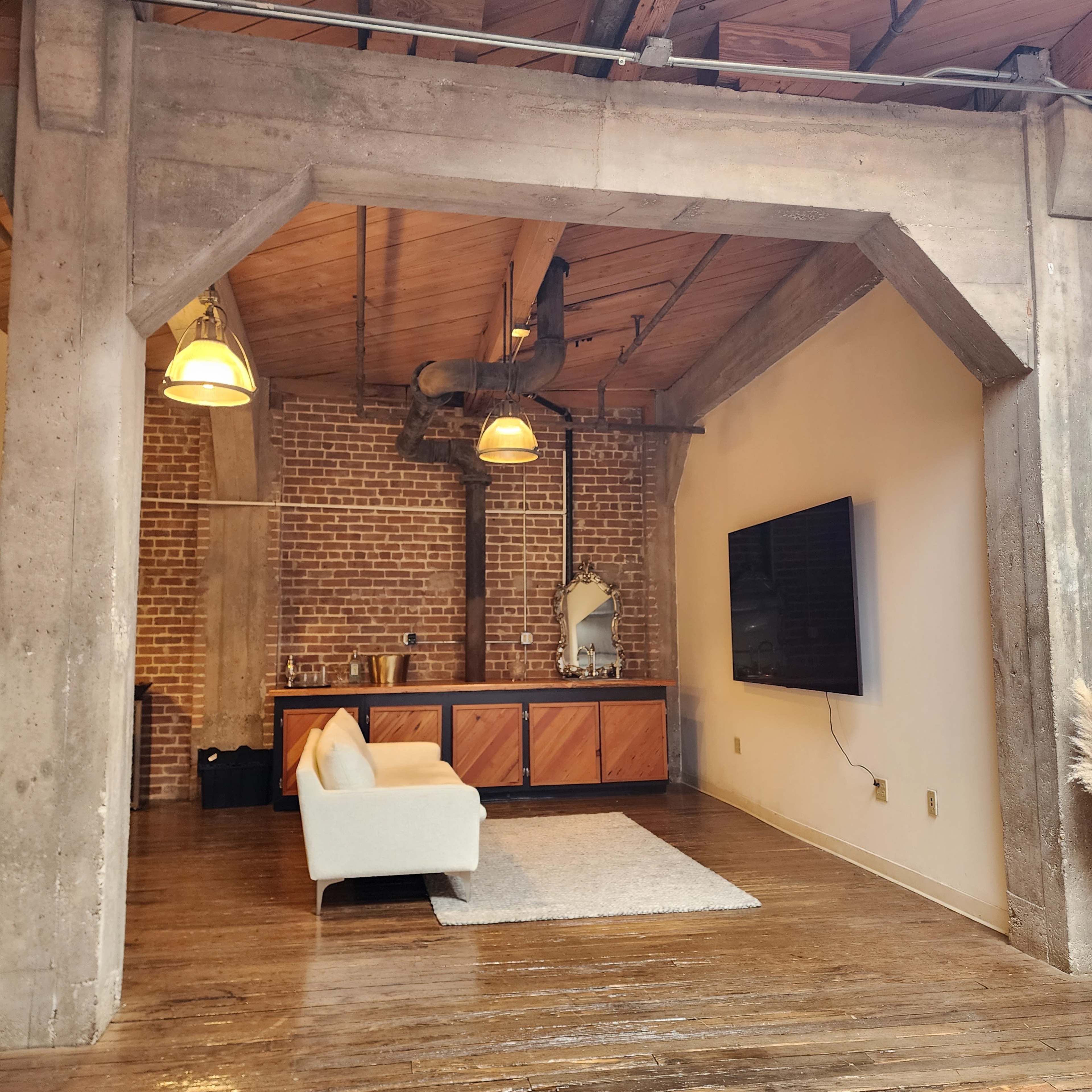 A spacious living area with exposed brick walls, a wooden ceiling, a modern sofa, a flat-screen TV mounted on the wall, and a wooden cabinet.