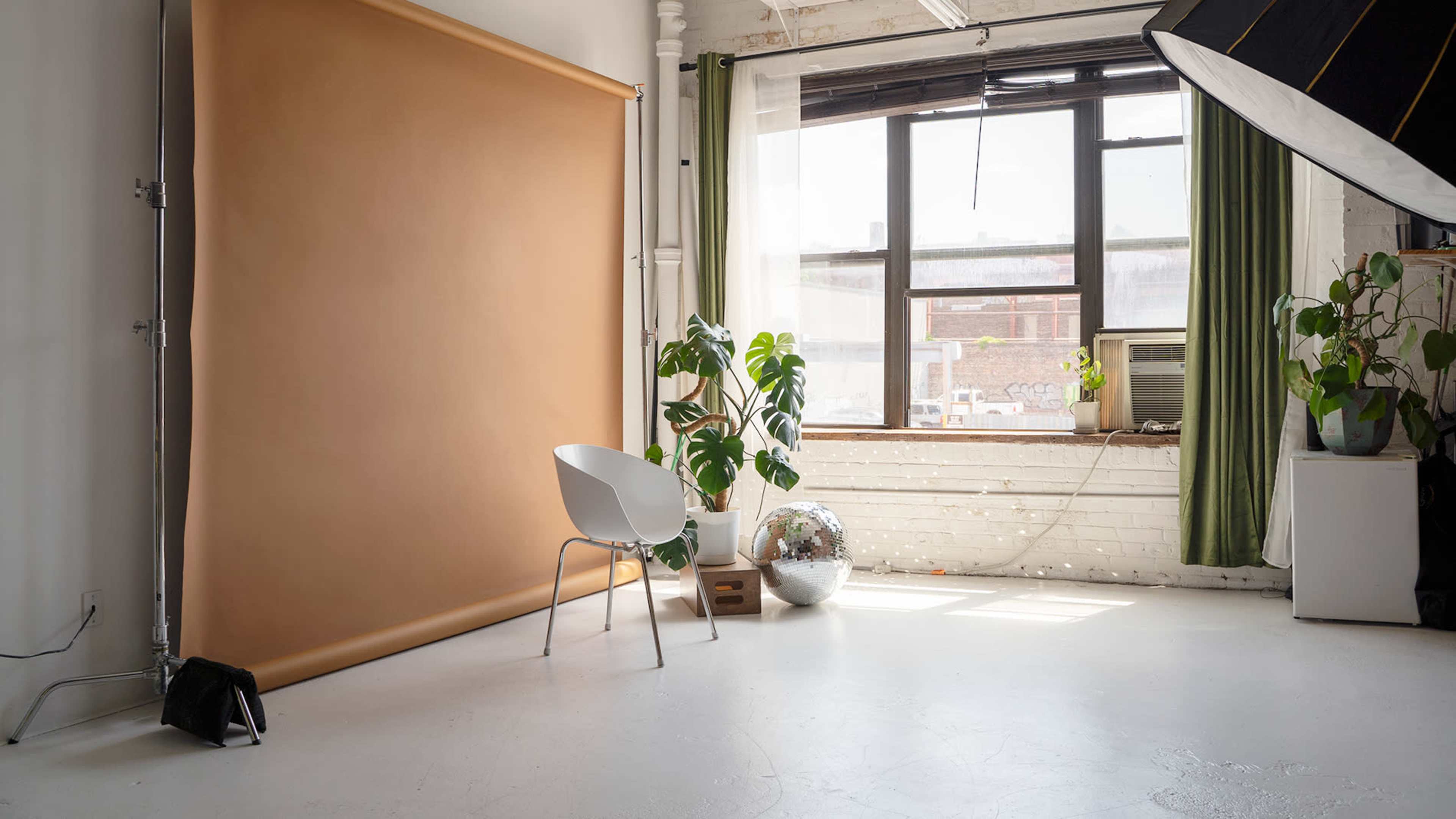 A photography studio featuring a light brown backdrop, a white chair, a large window with green curtains, and potted plants.