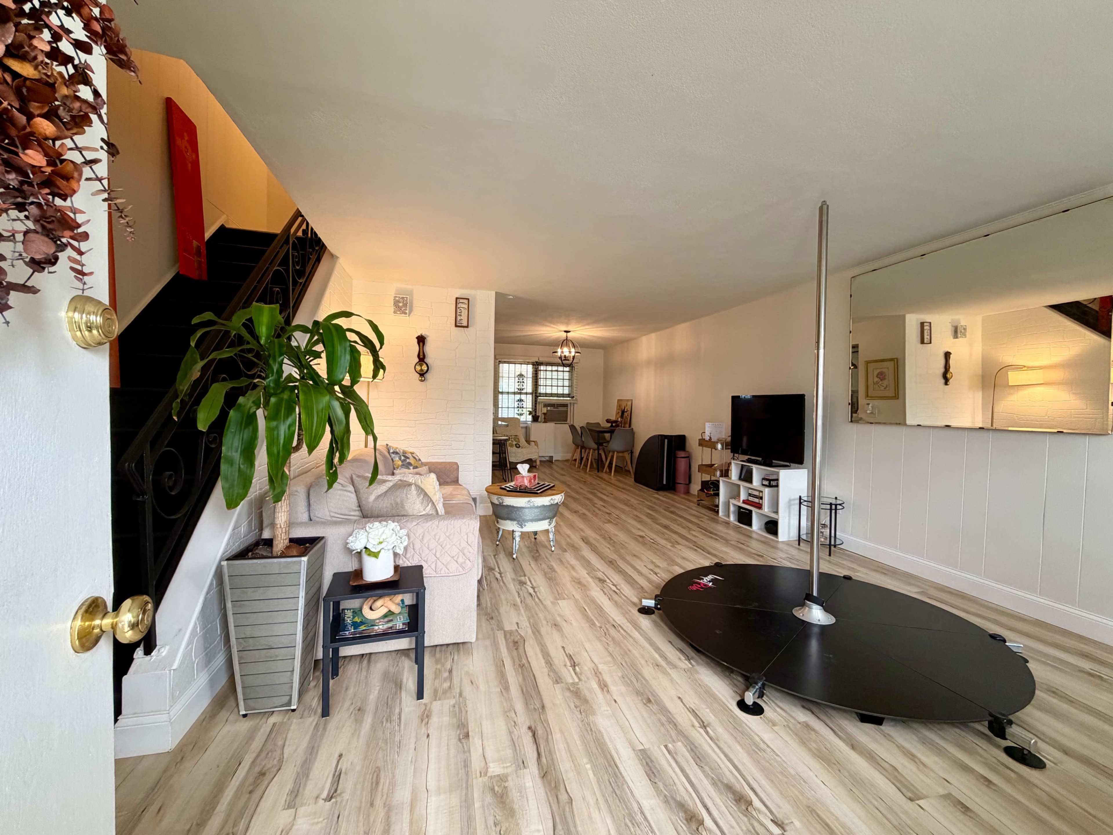 The image shows a modern living room with a pole dancer's pole, a sofa, a television, and a dining area, all complemented by wooden flooring and a staircase in the background.