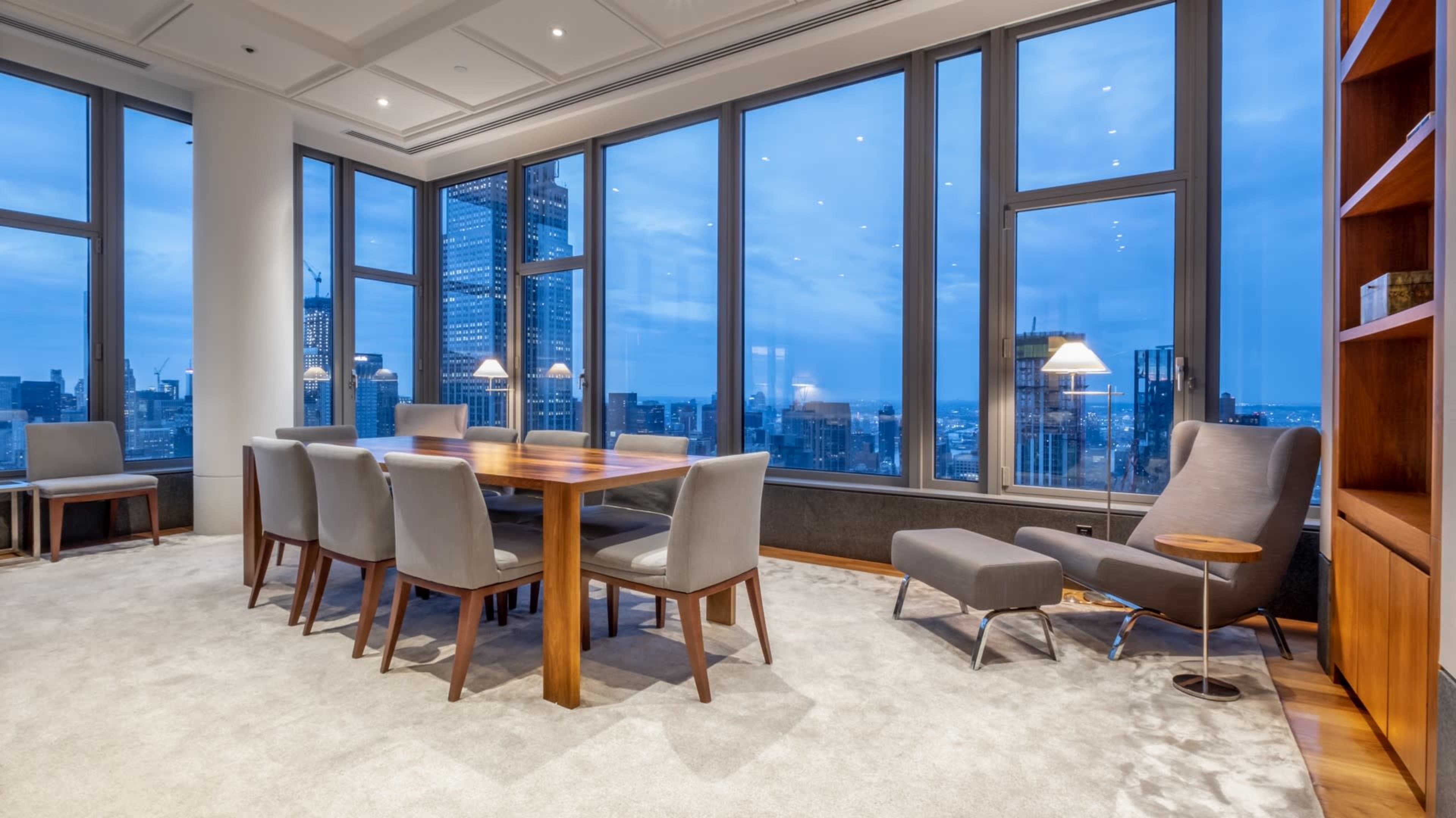 A modern conference room features a wooden table surrounded by gray chairs, with large windows showcasing a city skyline at twilight.
