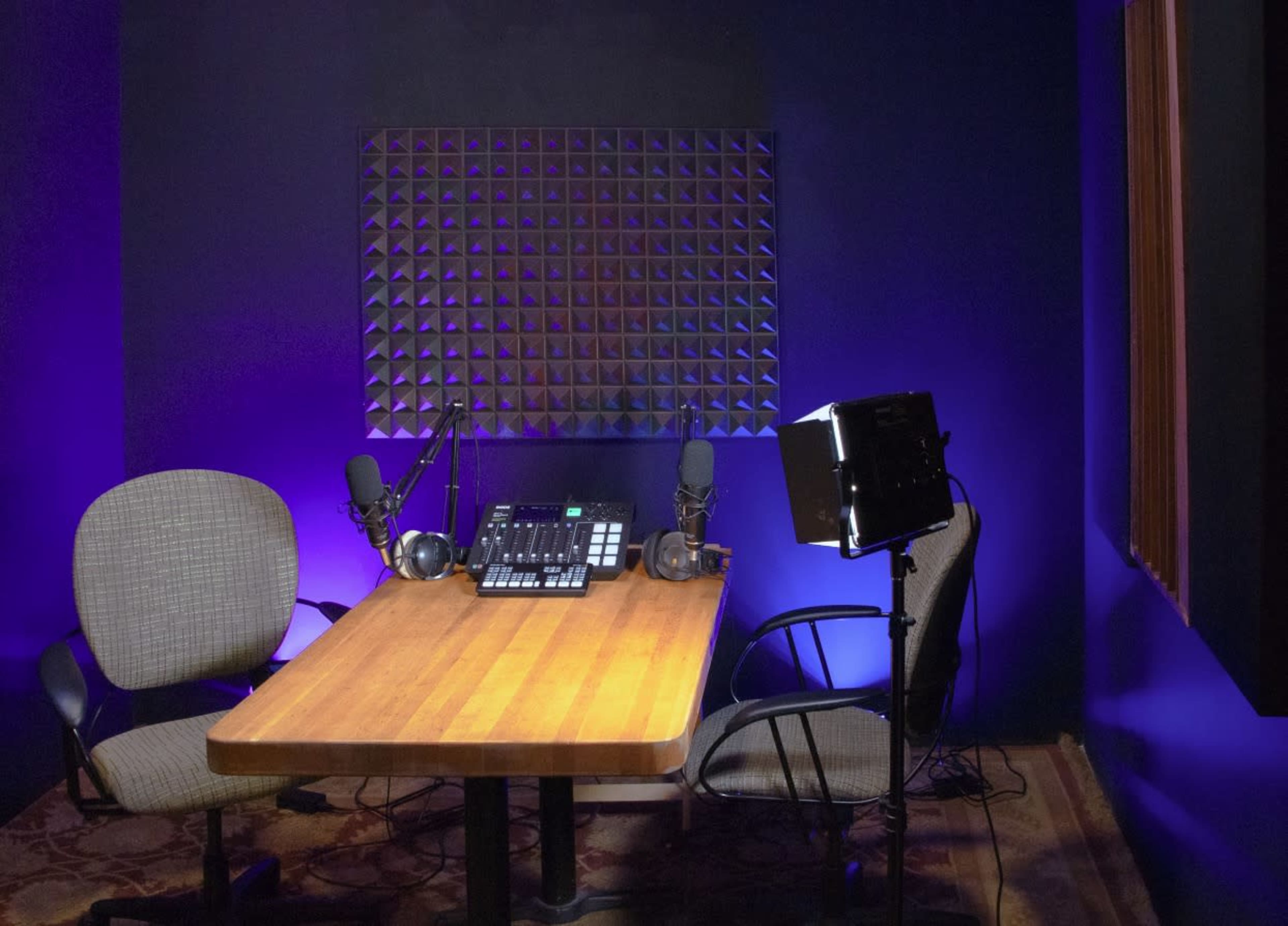 A podcast recording studio features two chairs, a wooden table, microphones, and lighting against a blue wall.