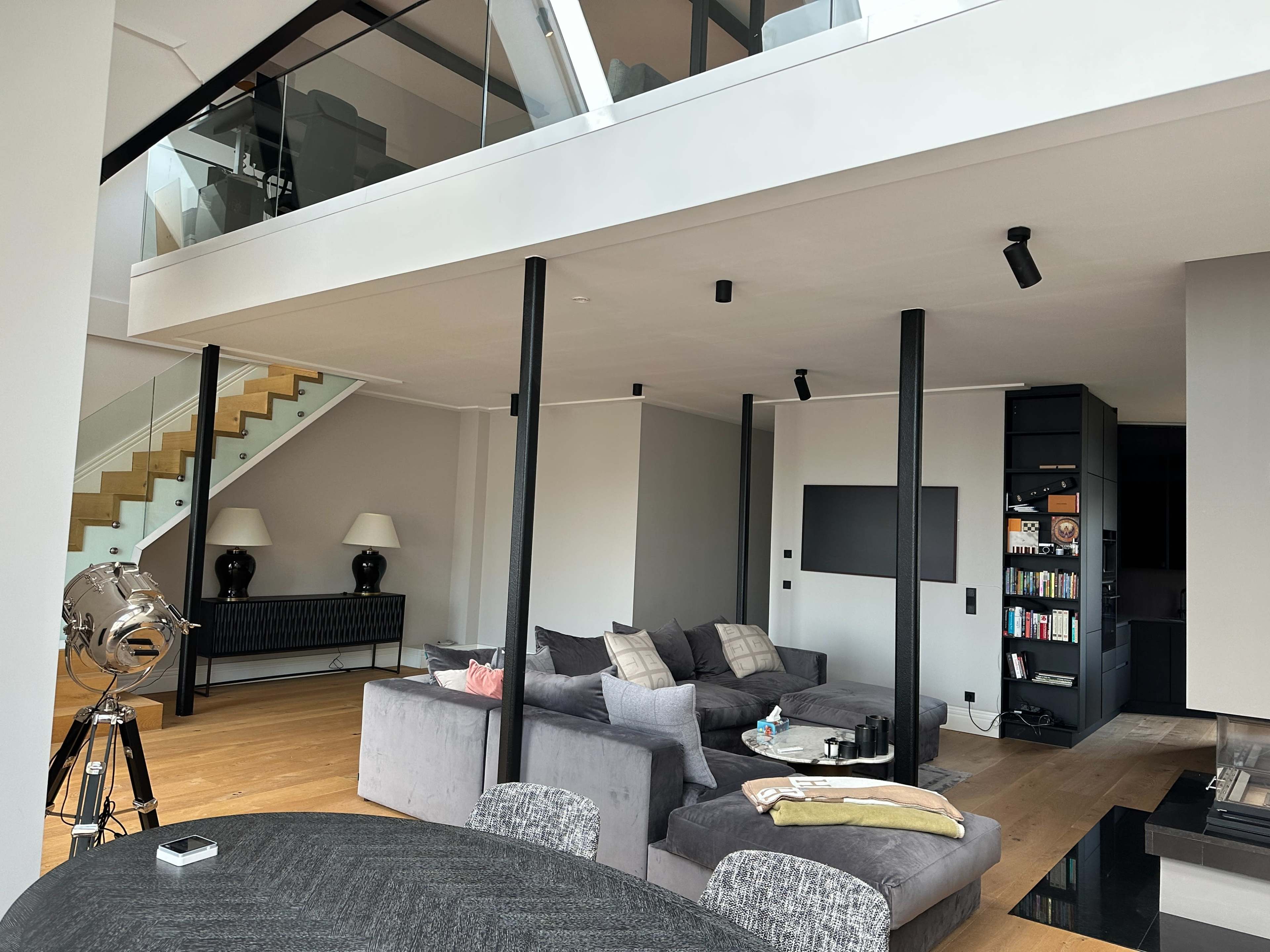 The image shows a spacious modern living room with a sectional sofa, a dining area, a television mounted on the wall, and a staircase leading to an upper level.