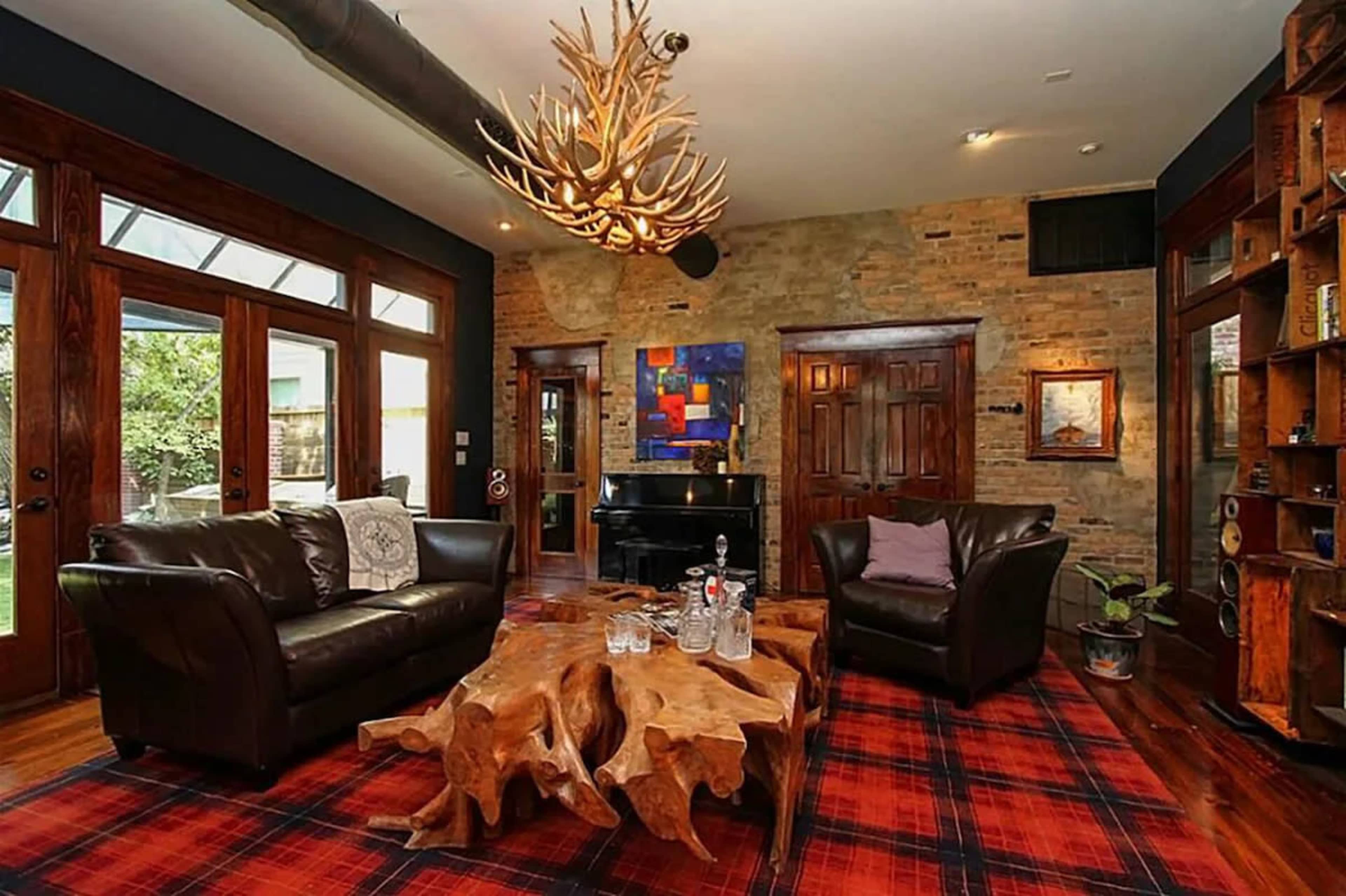 The living room features dark leather sofas, a wooden coffee table with a natural edge, and a grand piano against a wall of exposed brick and large windows.