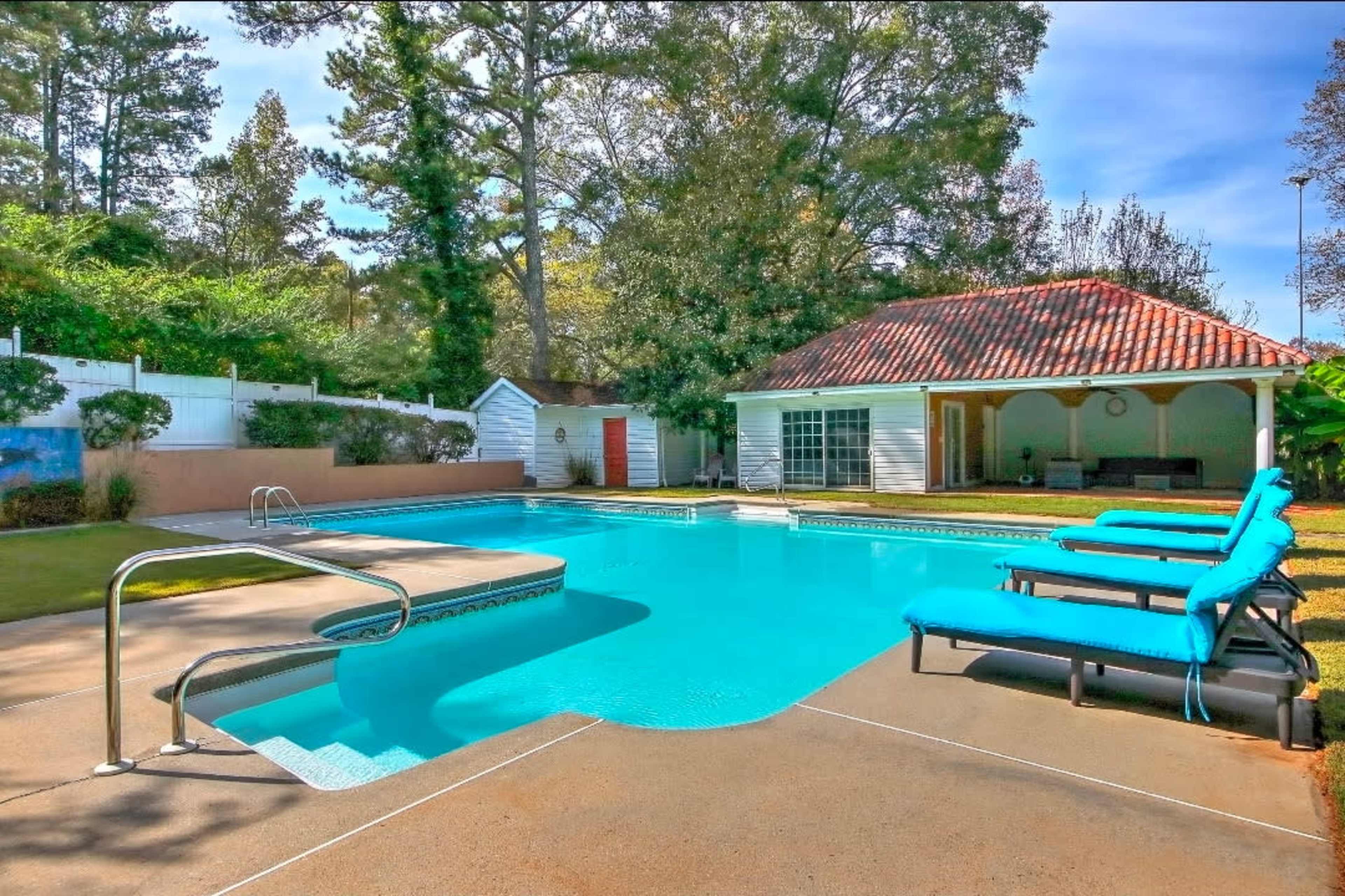 The image shows a clear swimming pool with blue lounge chairs beside it, adjacent to a white building with a red-tiled roof and surrounded by trees.
