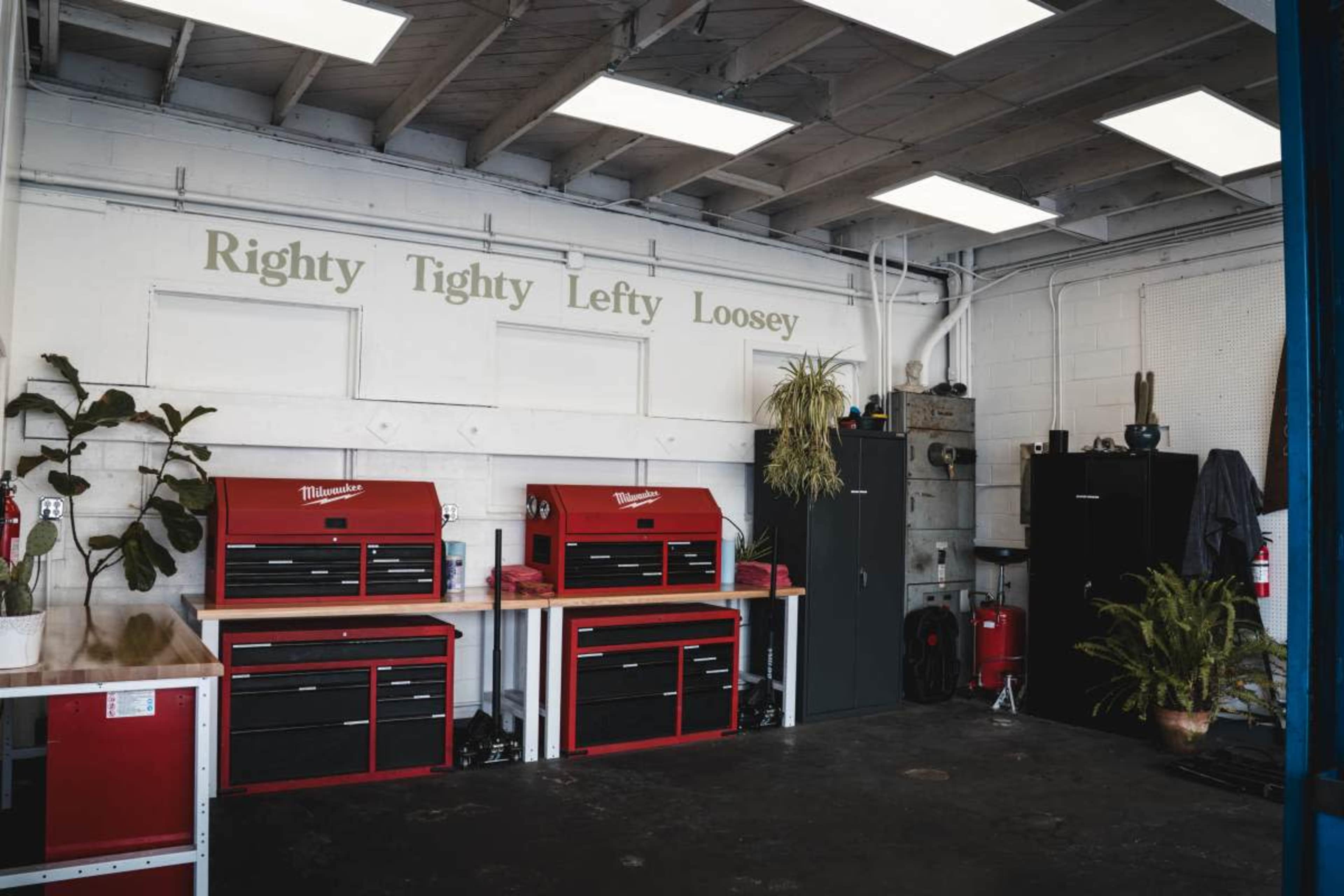 The image shows a clean and organized workshop with two red tool chests on white tables, a large black cabinet, and a plant against the wall featuring the phrase "Righty Tighty Lefty Loosey" above them.