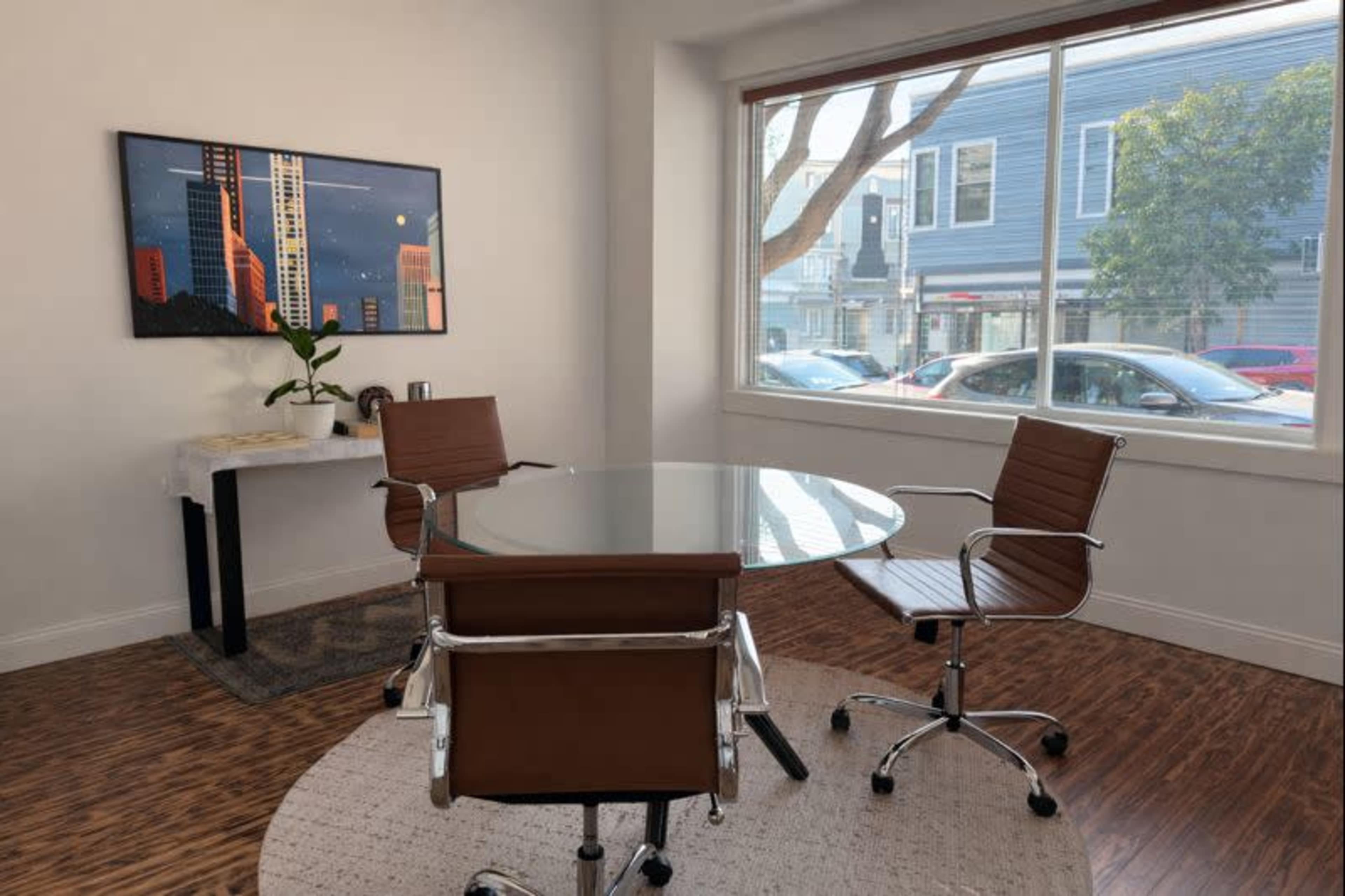 A modern office space features a round glass table surrounded by two brown leather chairs, with a plant and artwork on the walls, and a large window overlooking a street.