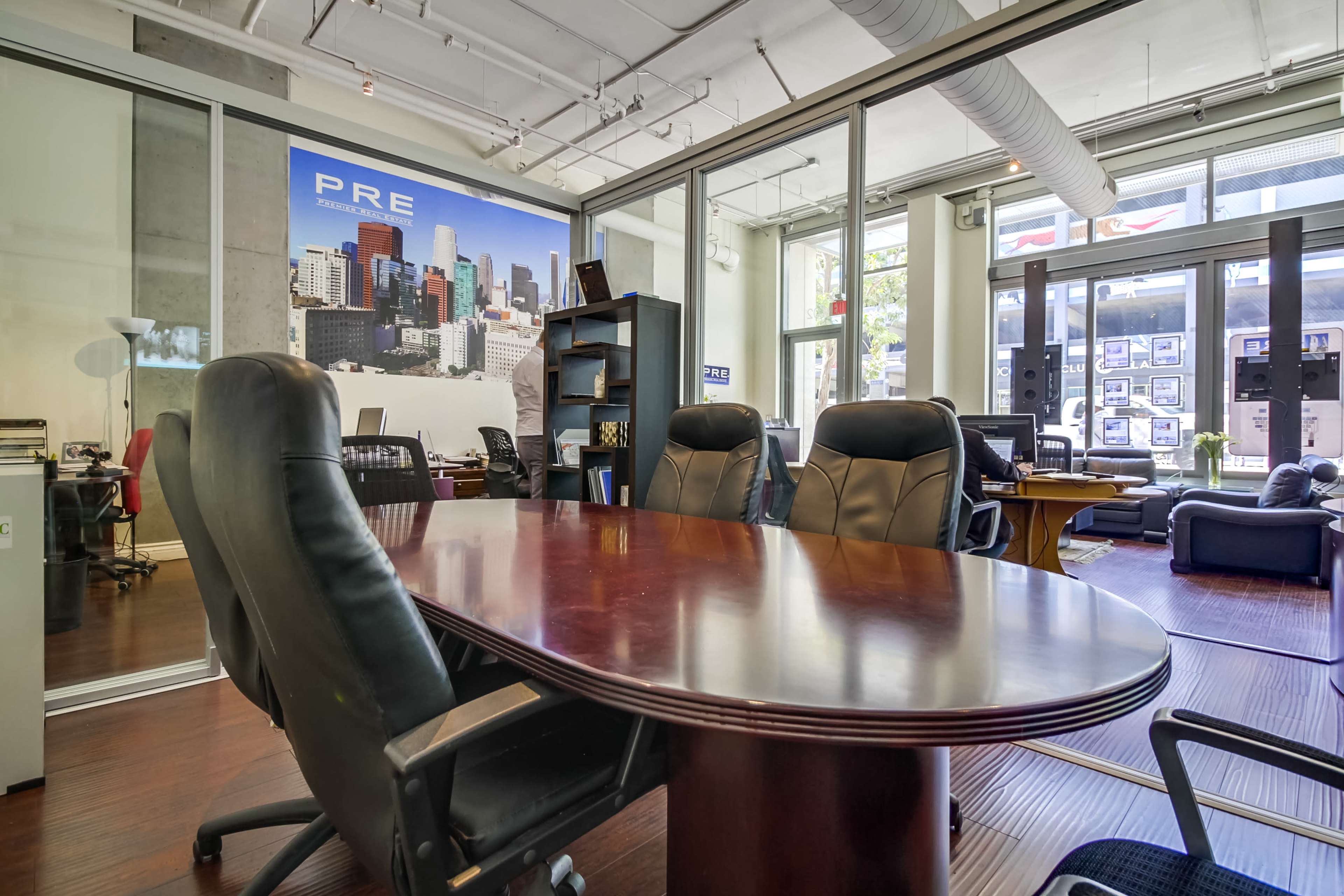 A conference room with a round wooden table and black leather chairs is visible, situated within an office space featuring large windows and a city skyline mural.