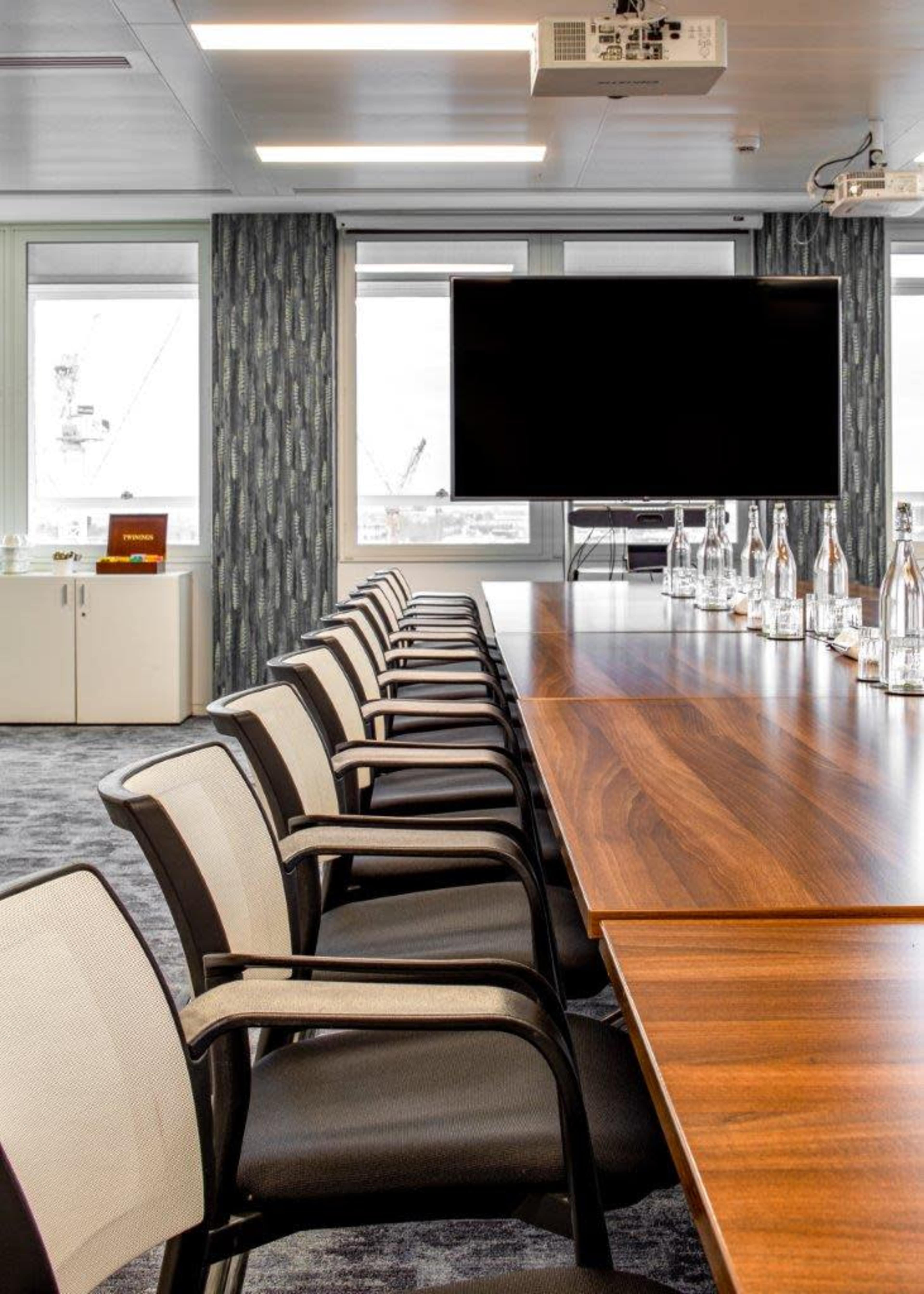 A long conference table with several chairs is set up in a modern meeting room featuring large windows and a wall-mounted screen.