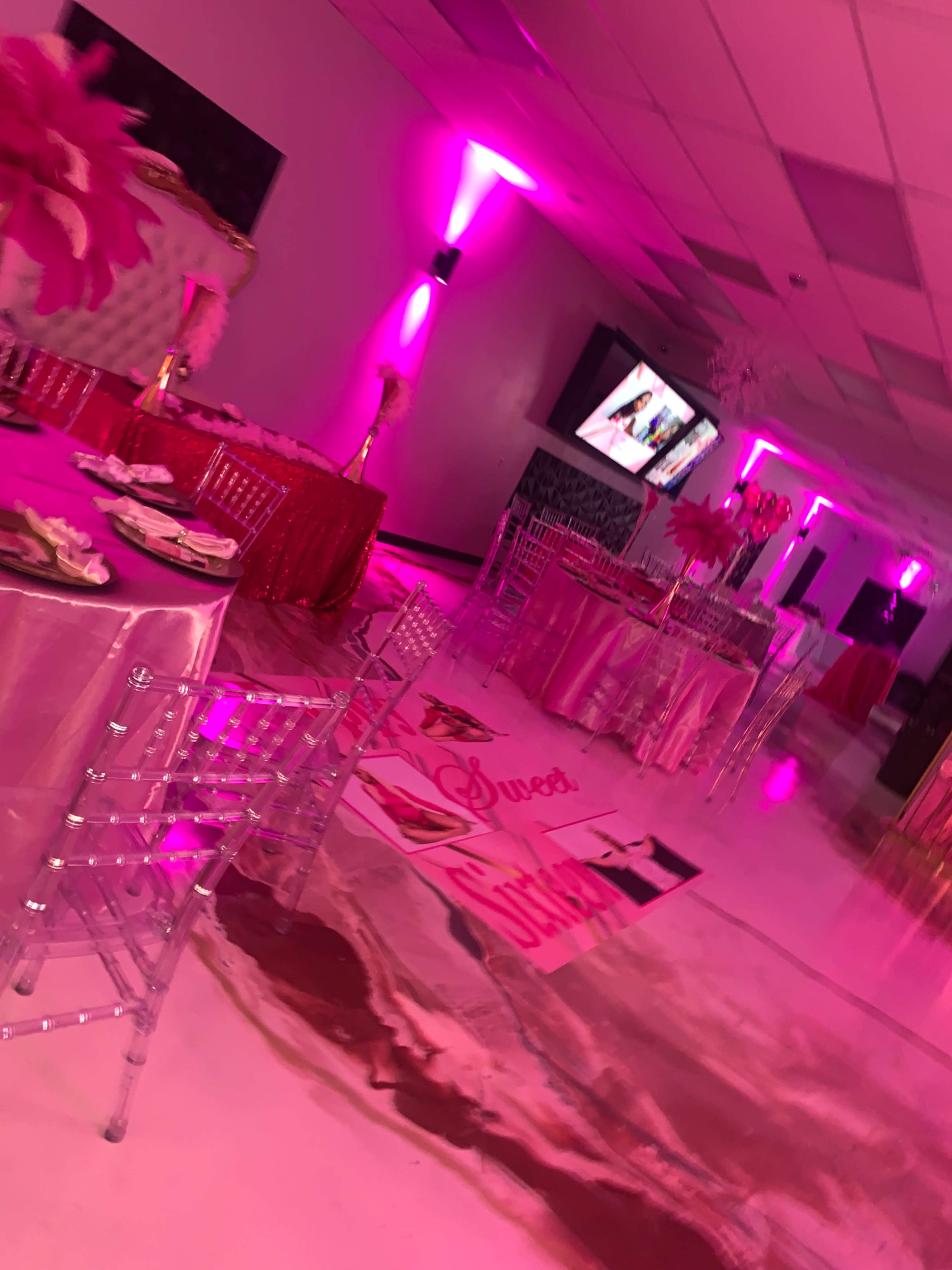 An event space decorated with pink lighting and table setups featuring transparent chairs and decorative linens.