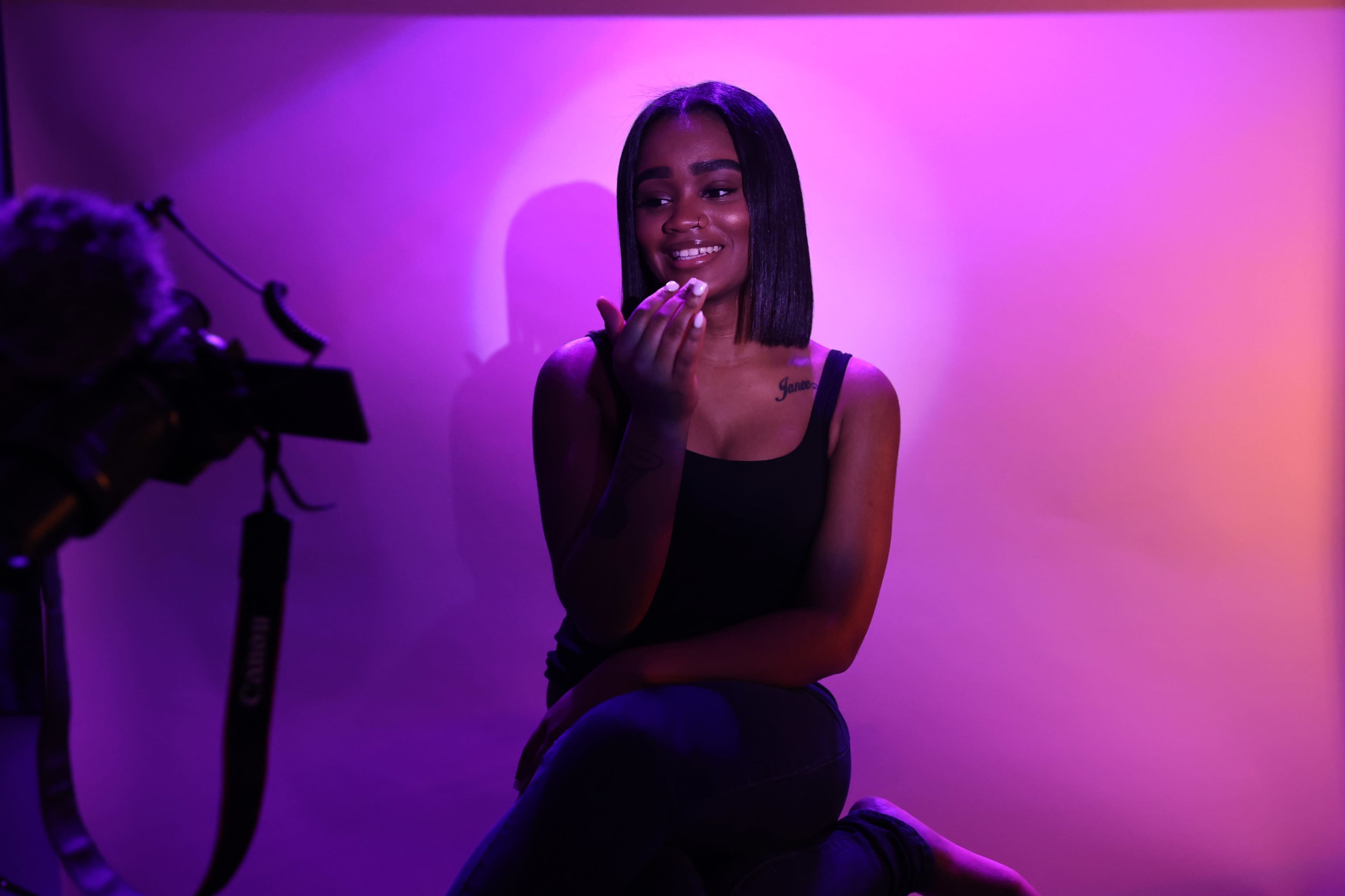 A woman sits on a stool in front of a camera, illuminated by purple background lighting.