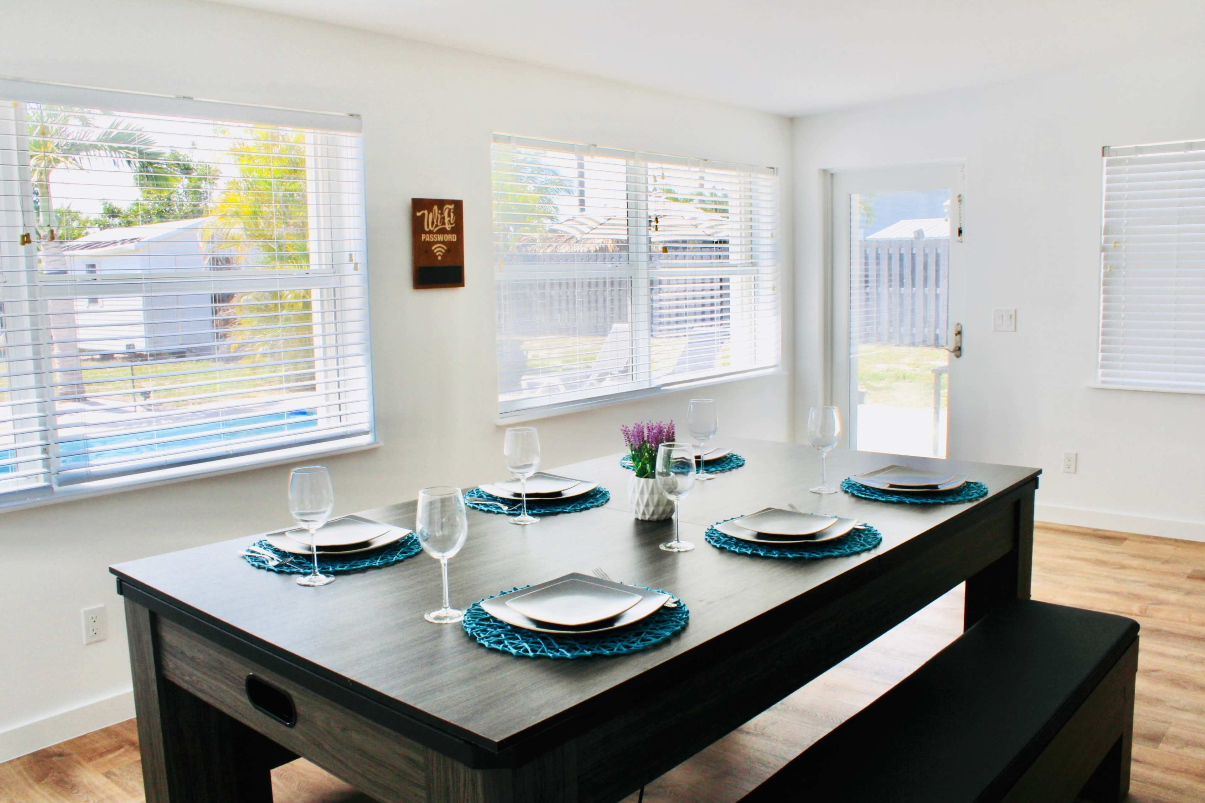 A modern dining room features a rectangular table set with plates, glasses, and decorative placemats, surrounded by large windows that provide ample natural light.