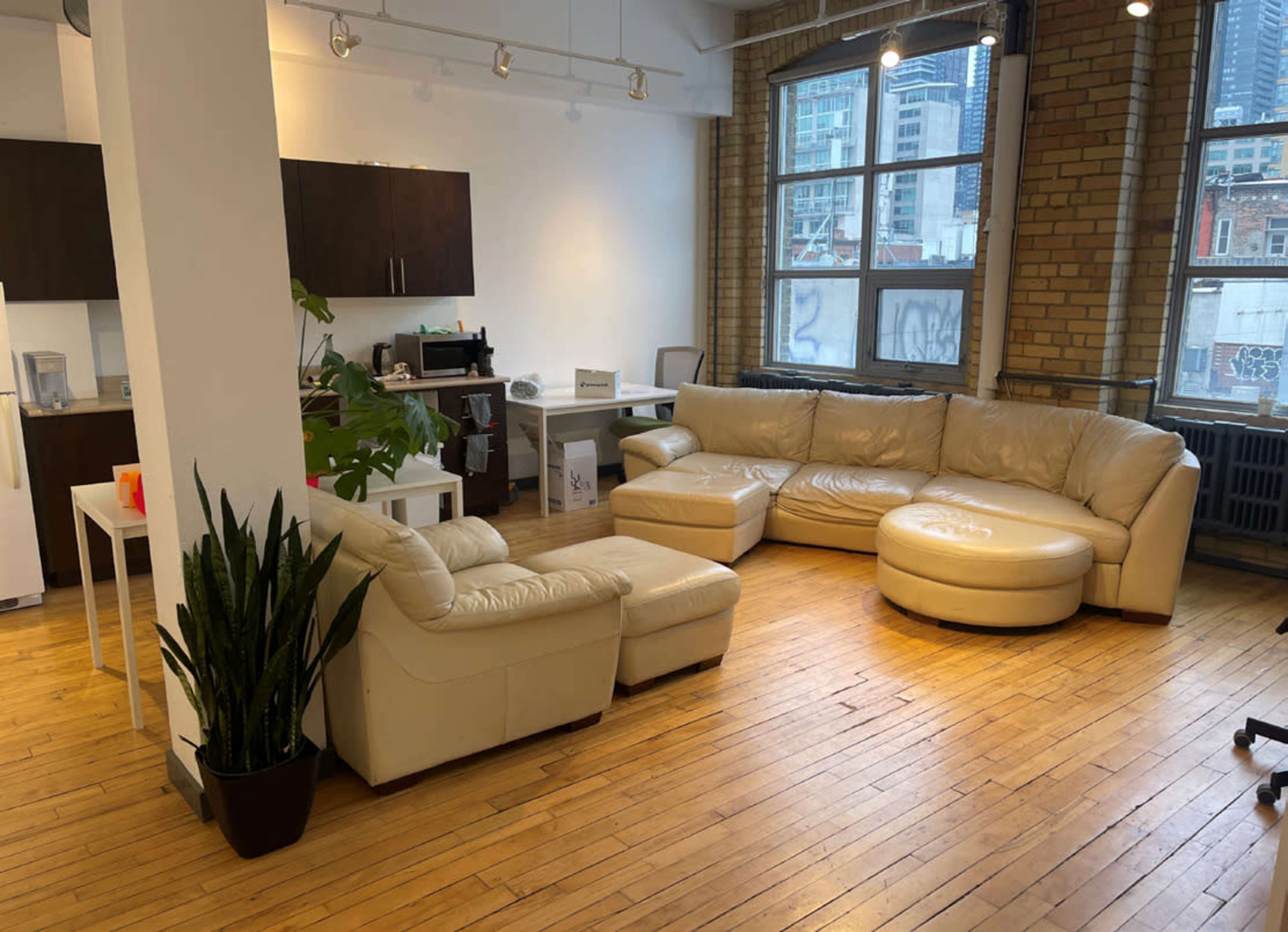 A spacious room with a large beige leather sectional sofa, a white desk, and large windows overlooking a cityscape.