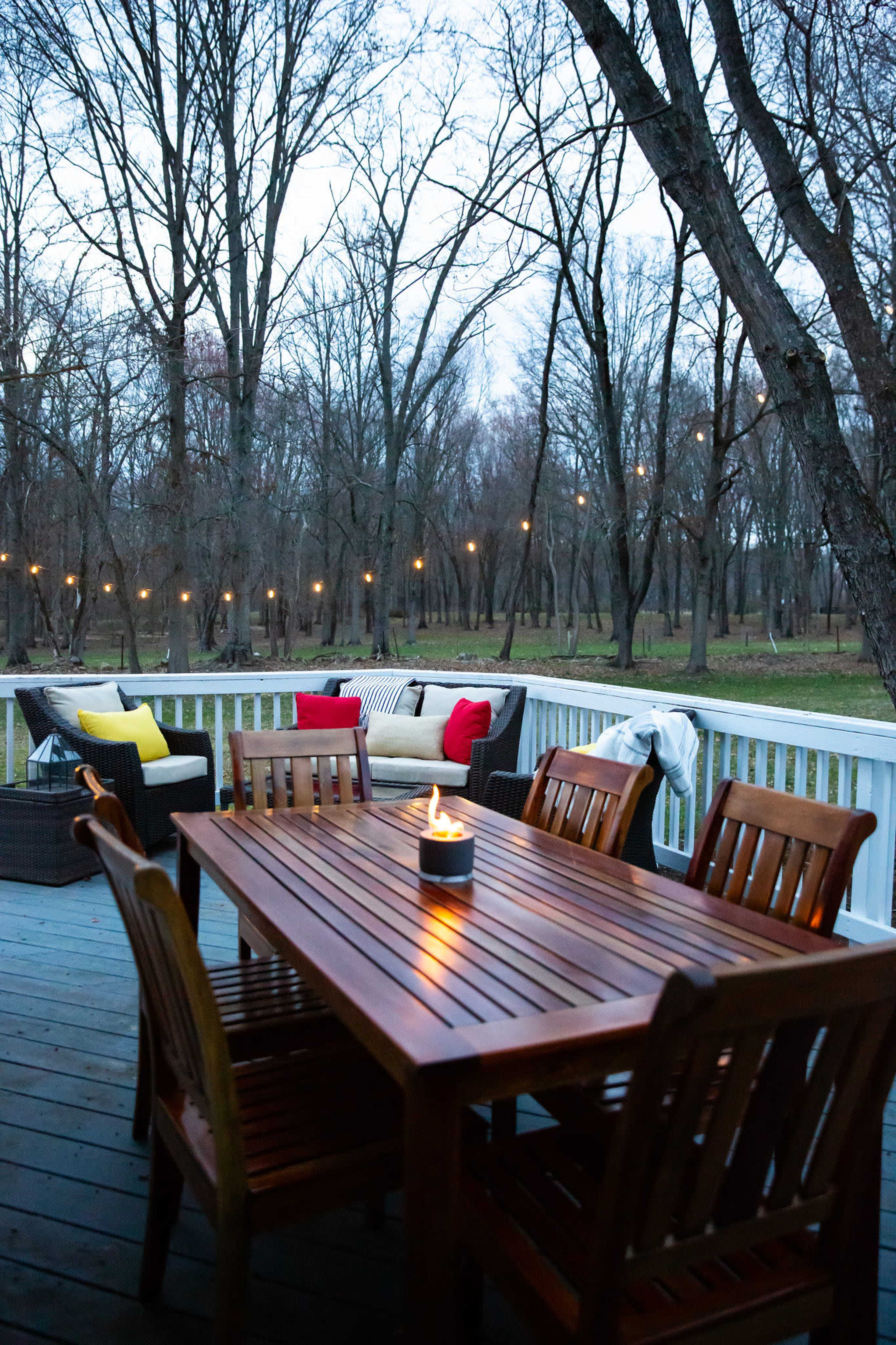A wooden deck features a dining table with chairs and a small fire centerpiece, surrounded by trees in a twilight setting.