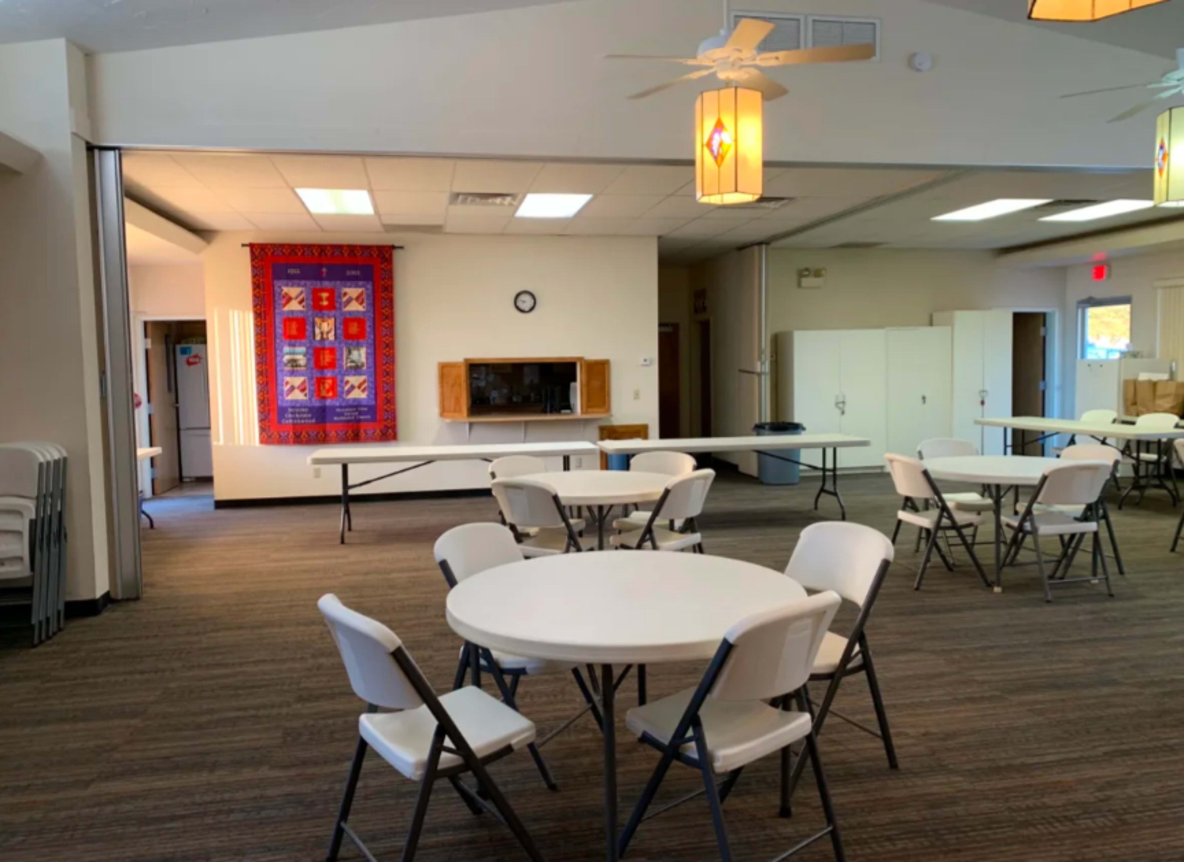 A spacious community hall with several circular tables and chairs arranged, and a colorful quilt hanging on the wall in the background.