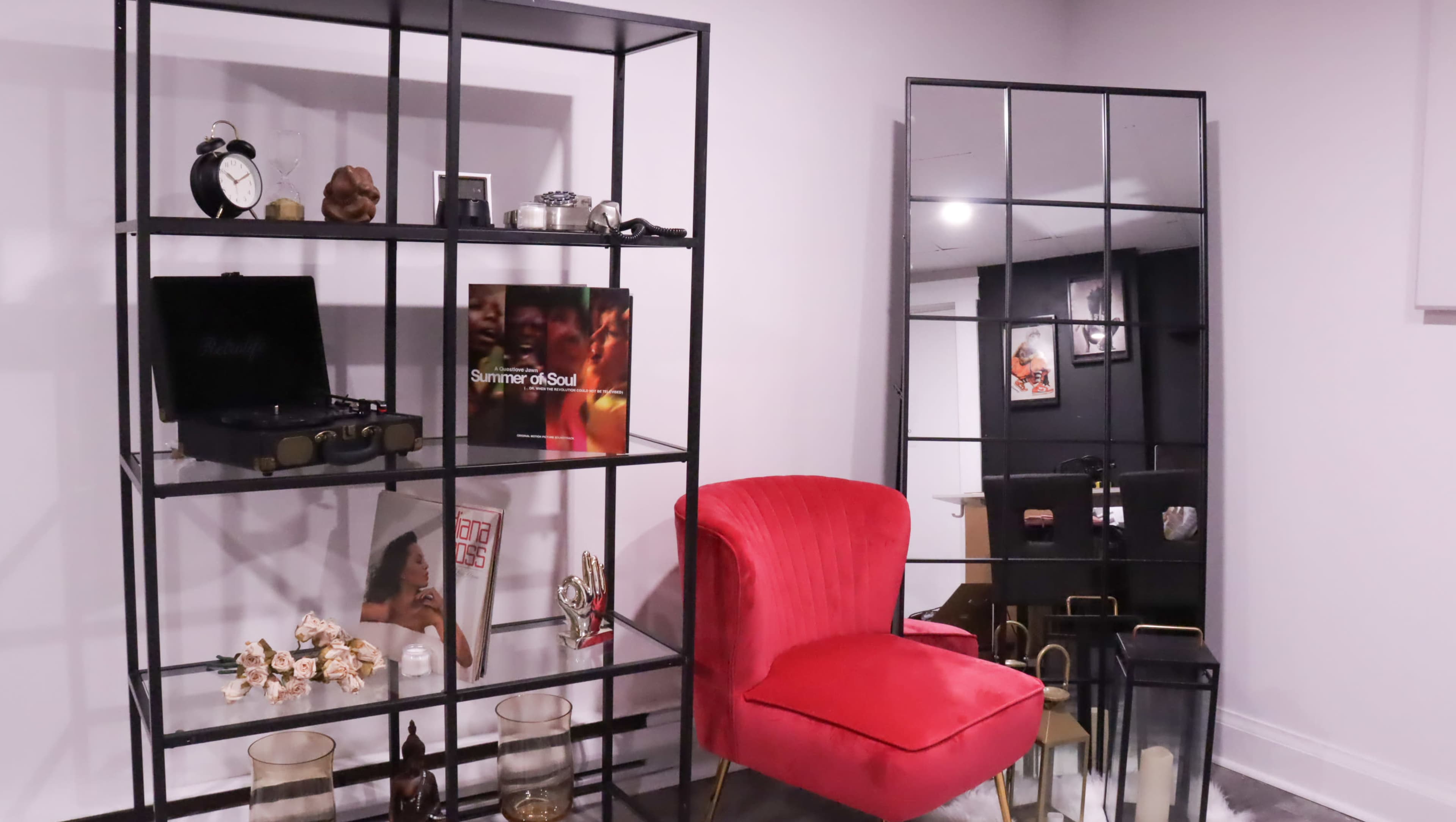 A modern room featuring a black bookshelf filled with decorative items, a red velvet chair, and a large mirror on the wall.