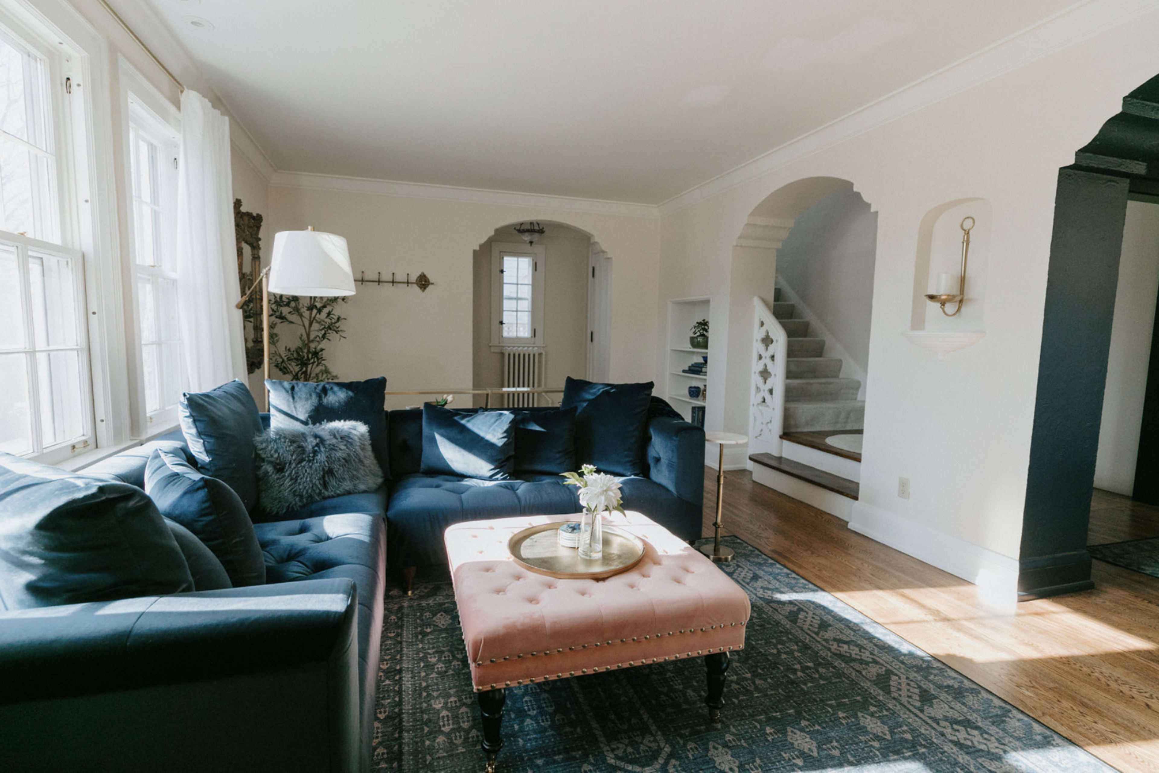 A blue sectional sofa and a pink ottoman are arranged in a bright living room with large windows and a view of a staircase.