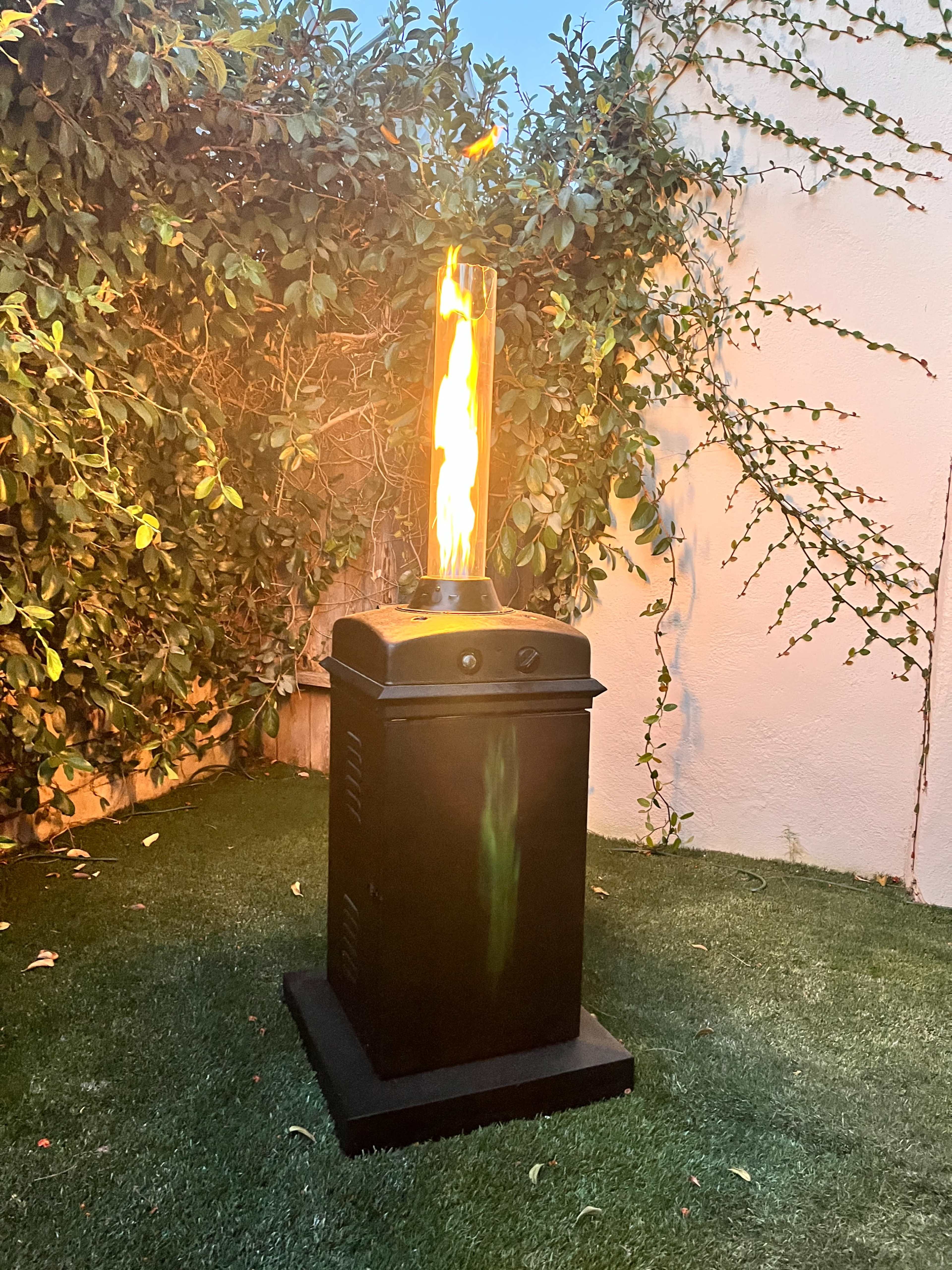A tall gas flame feature stands on a grassy surface surrounded by greenery.