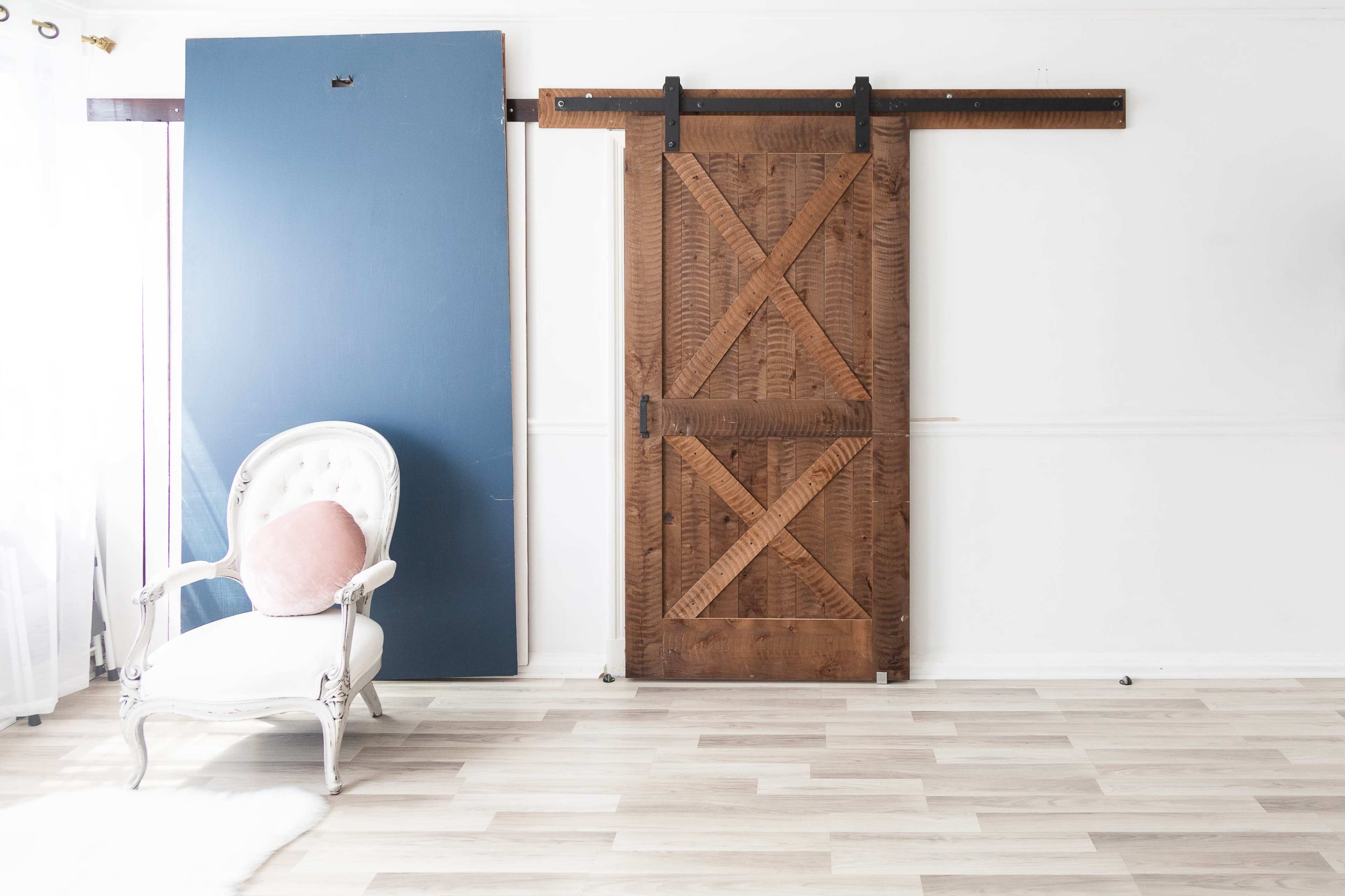 A wooden sliding barn door, a blue panel, and a white upholstered chair with a pink cushion positioned beside it in a well-lit room.