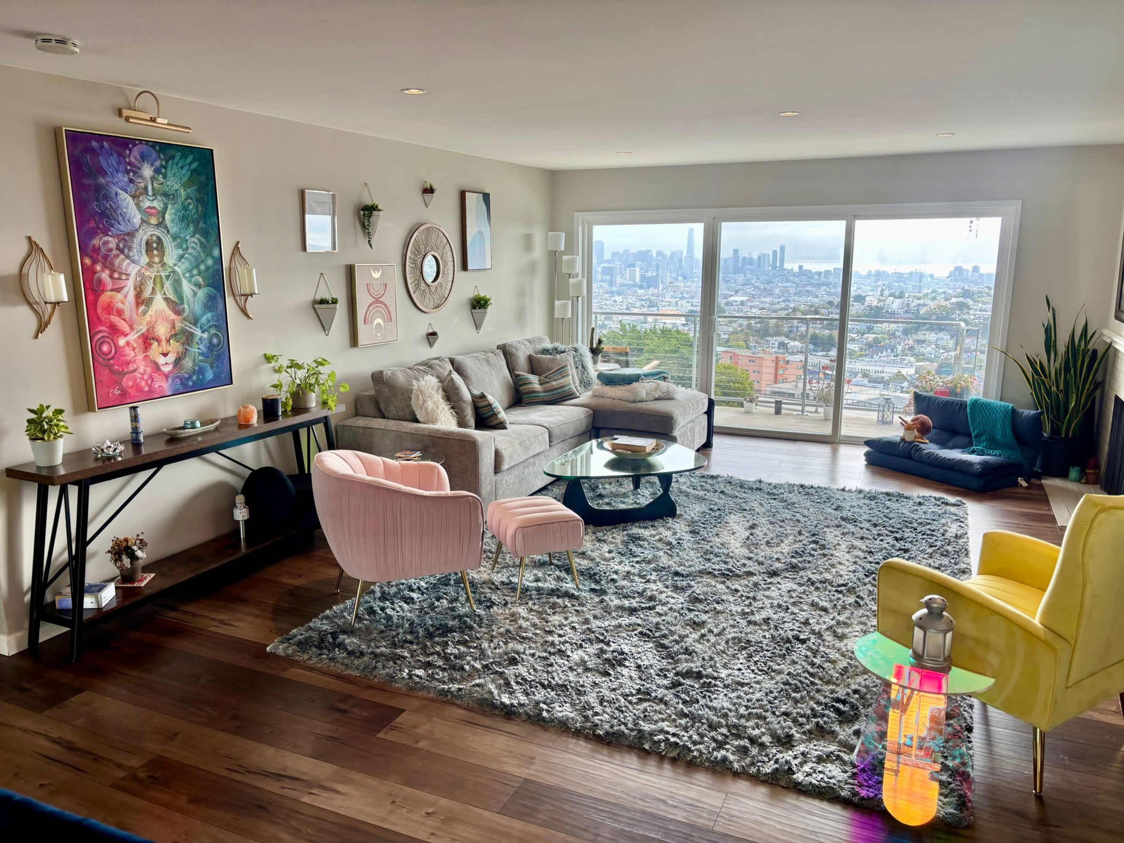 The image shows a modern living room with a large window offering a city view, featuring a gray sofa, a colorful rug, and various decorative plants and furniture.
