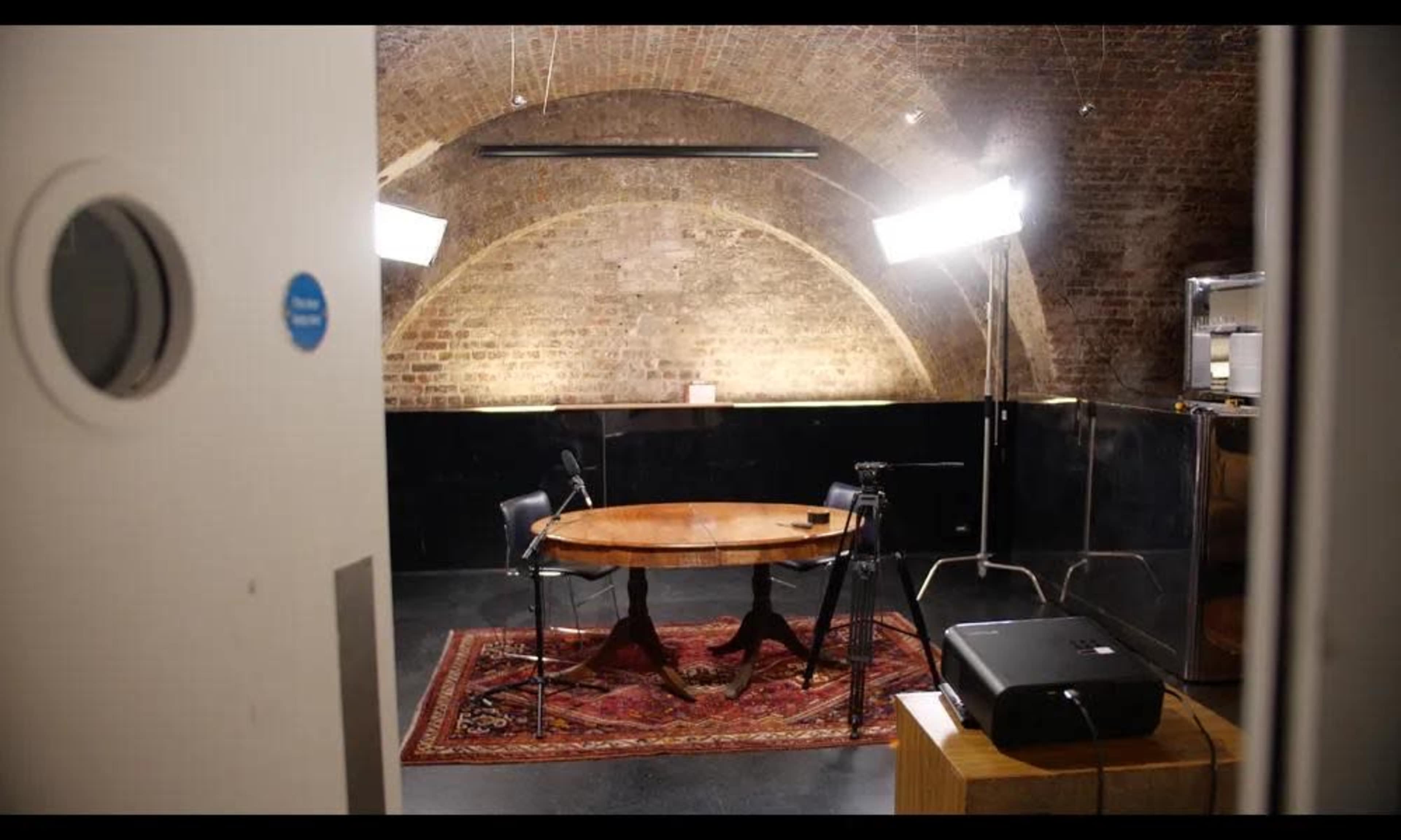 The image shows a small studio with a round table and two chairs, illuminated by soft lighting, set against a backdrop of exposed brick walls.