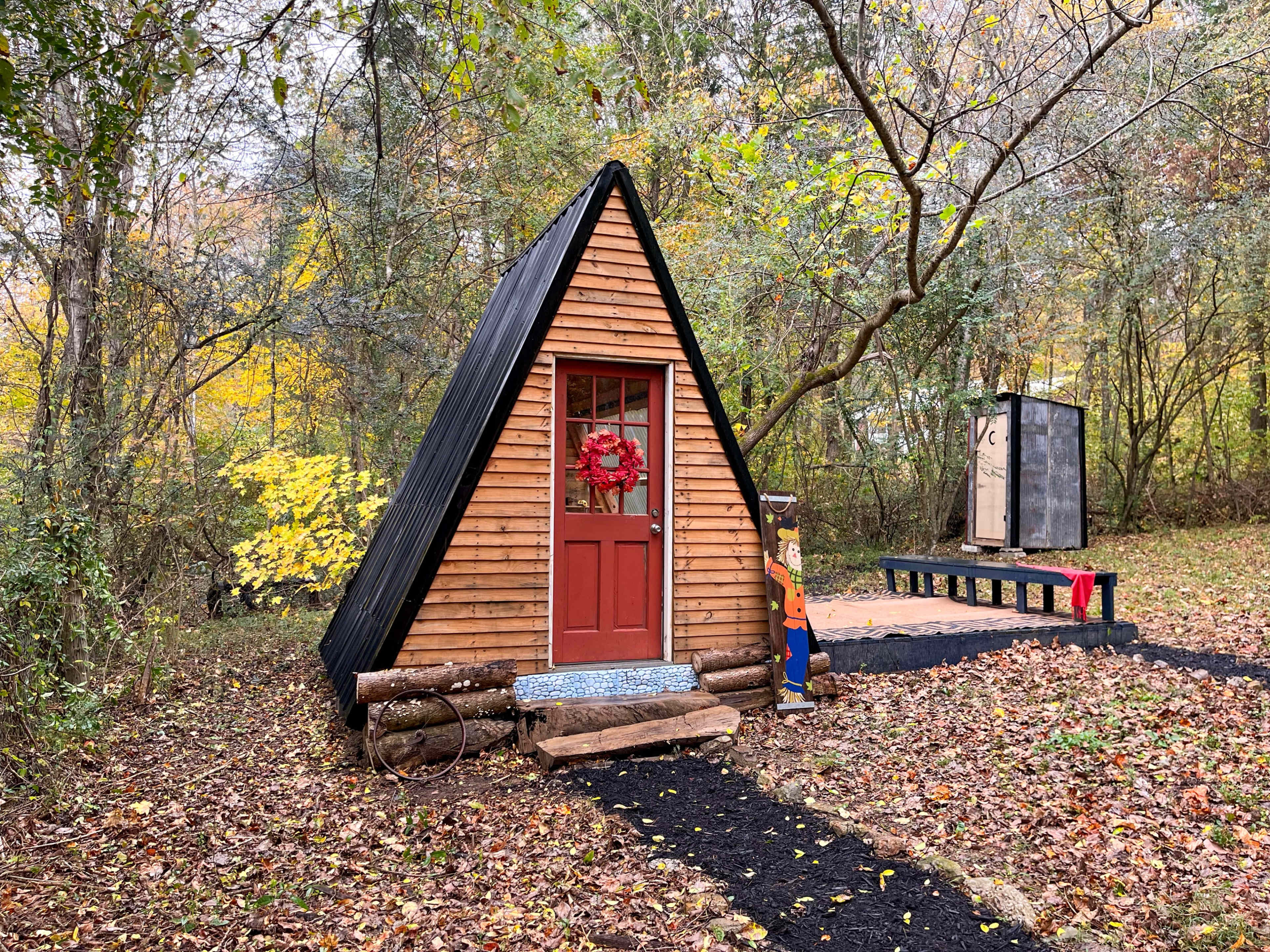 A small A-frame cabin with a red door and window, surrounded by autumn foliage and a path leading to a platform.