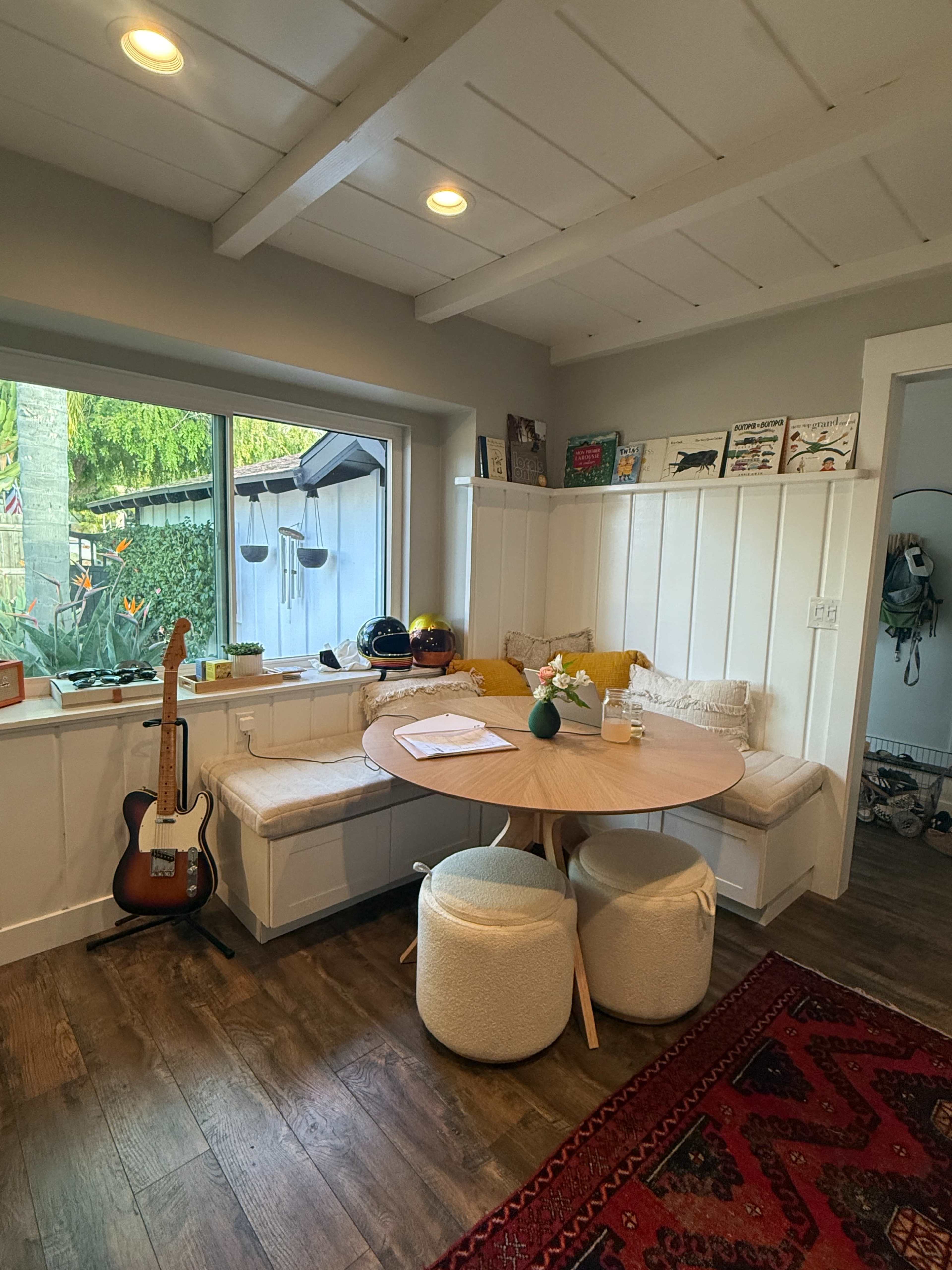 The image shows a cozy dining nook with a round wooden table surrounded by built-in seating, a guitar resting against the wall, and a window view of greenery outside.