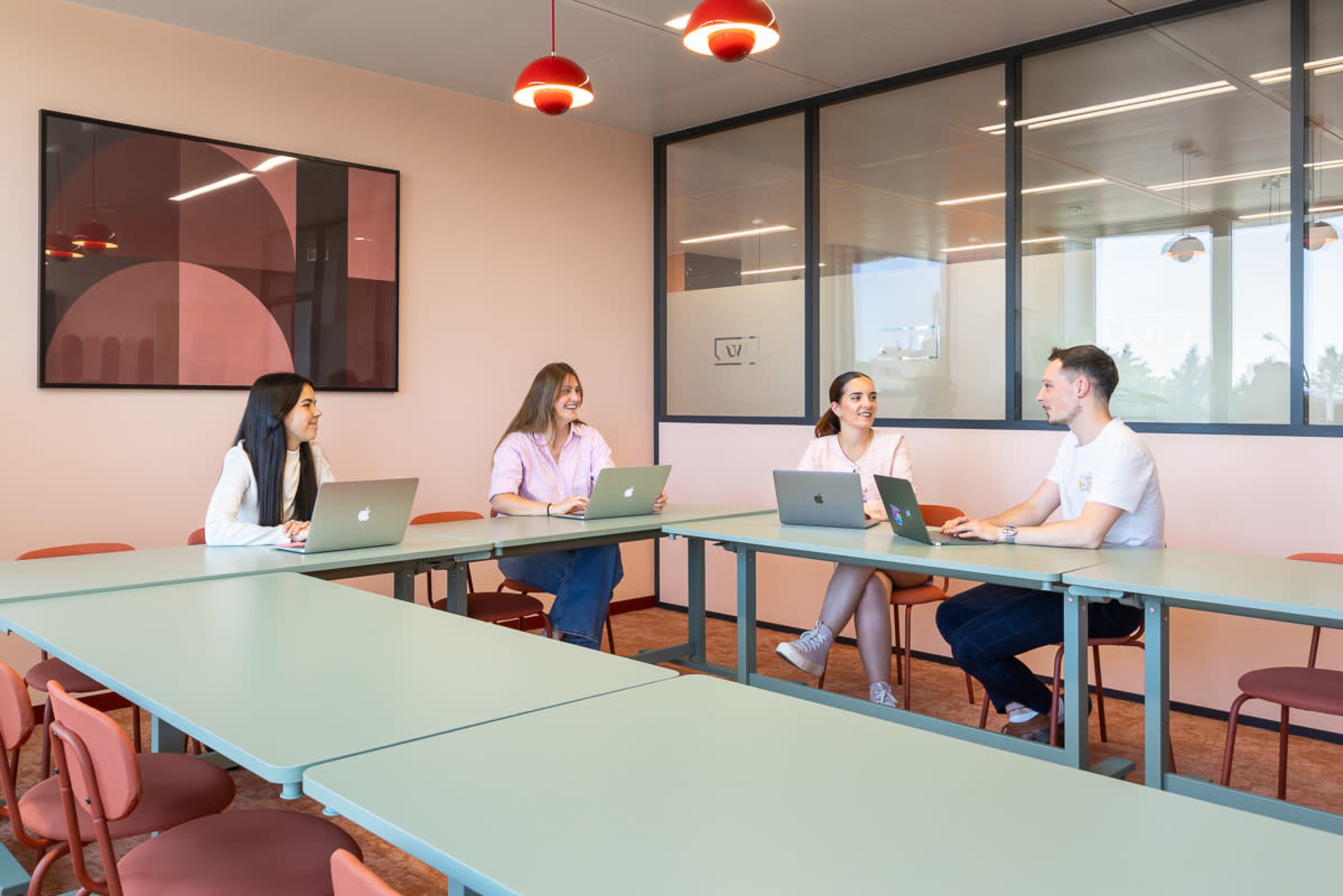 Four people are seated at a large table in a modern meeting room, each using a laptop.