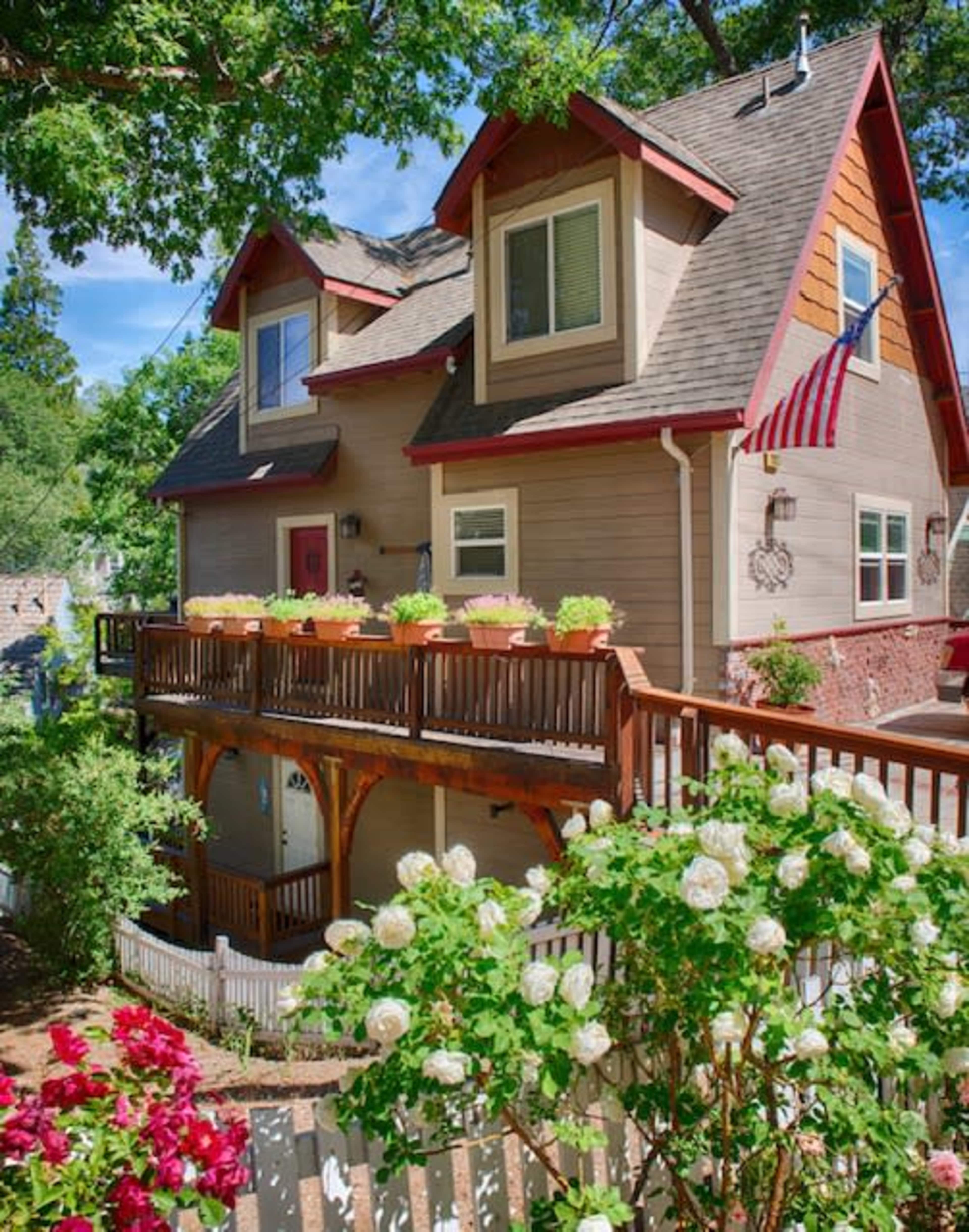 A two-story house with a wooden deck adorned with flower pots and a flag, surrounded by a garden featuring rose bushes and a white picket fence.