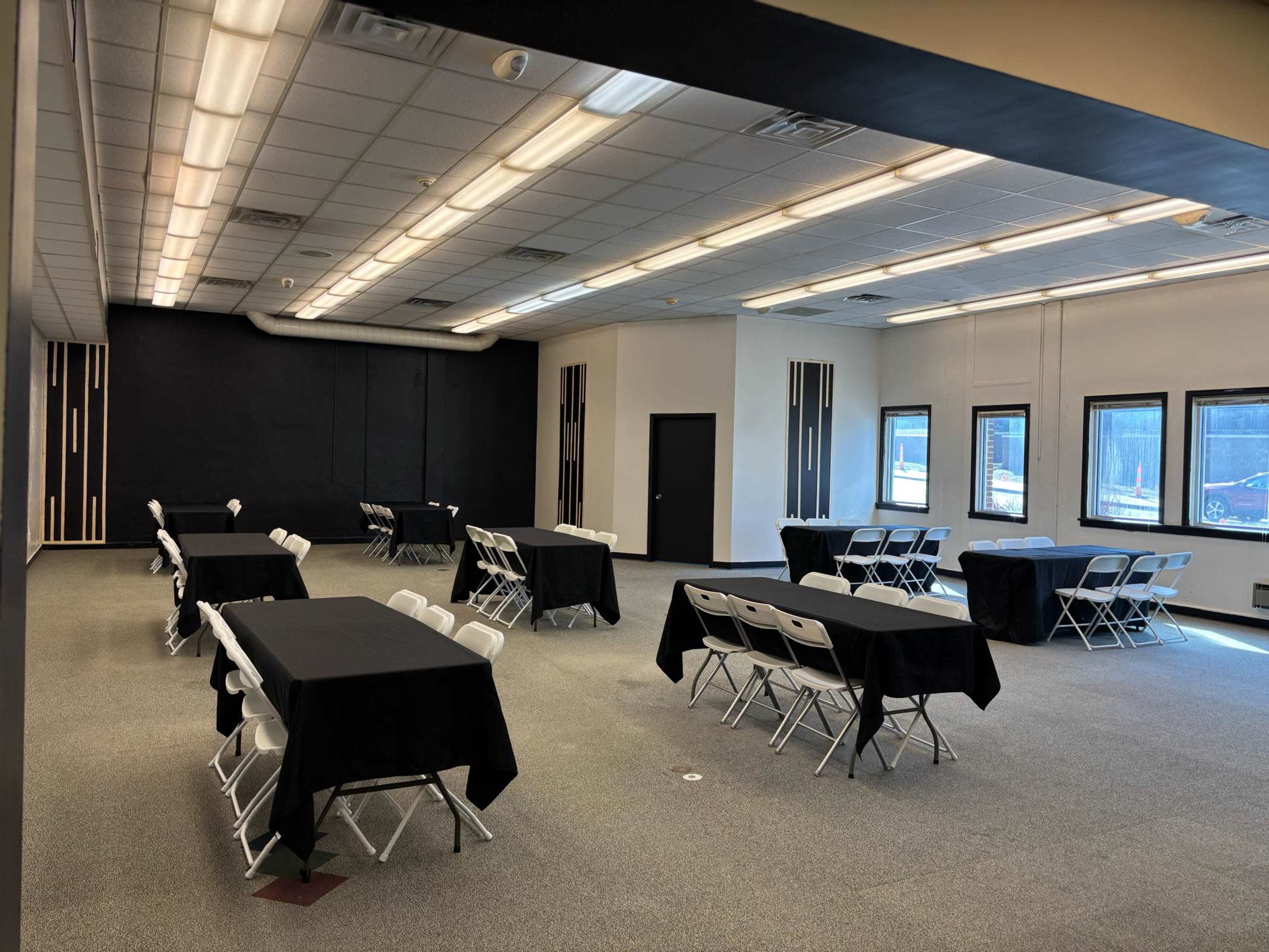 The image shows a spacious room set up with several tables covered in black cloth and surrounded by folding chairs, with large windows along one wall.
