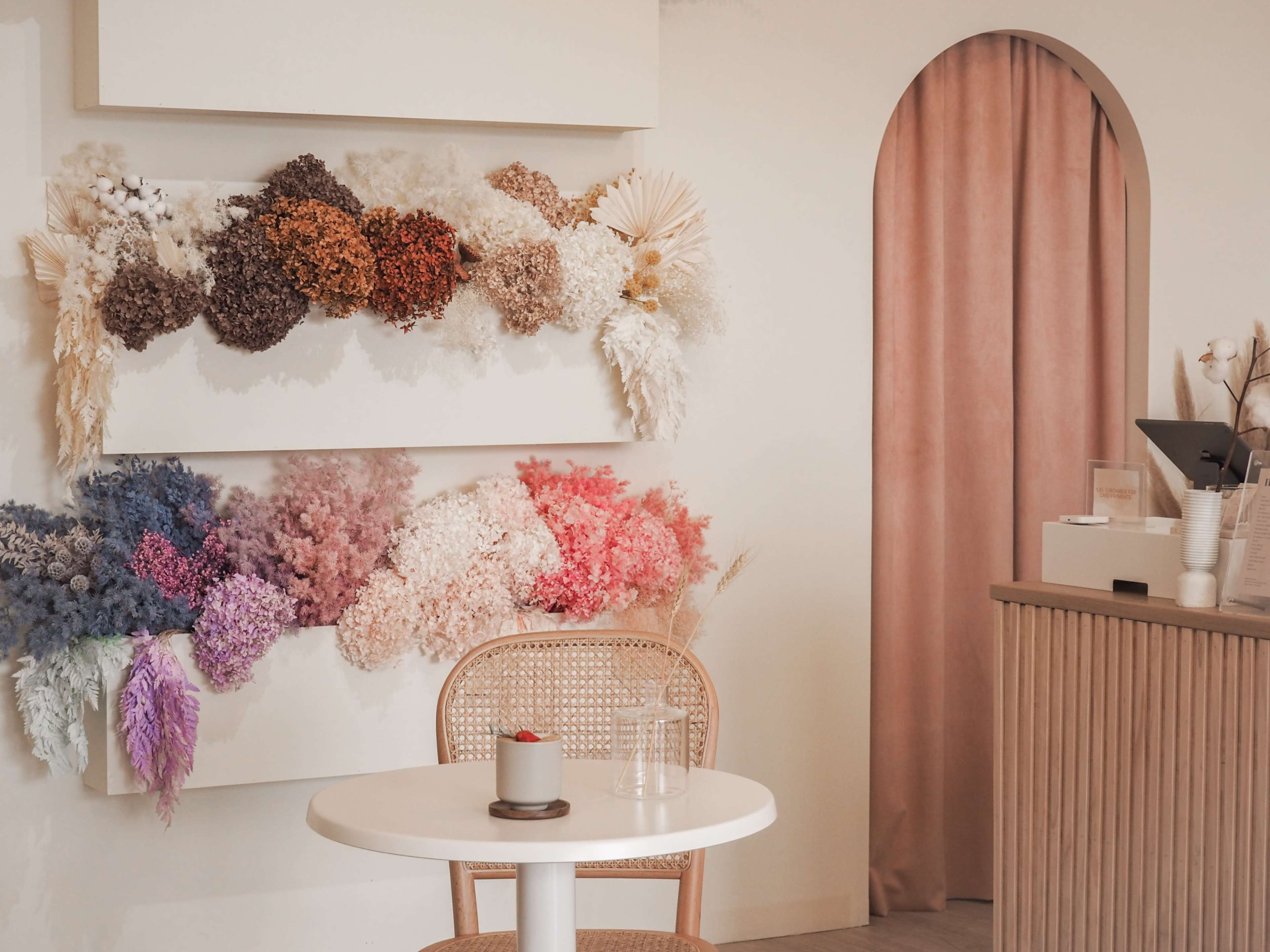 A minimalist interior features a white wall adorned with a variety of dried flowers, a round table with a small dessert, and a beige curtain in the background.