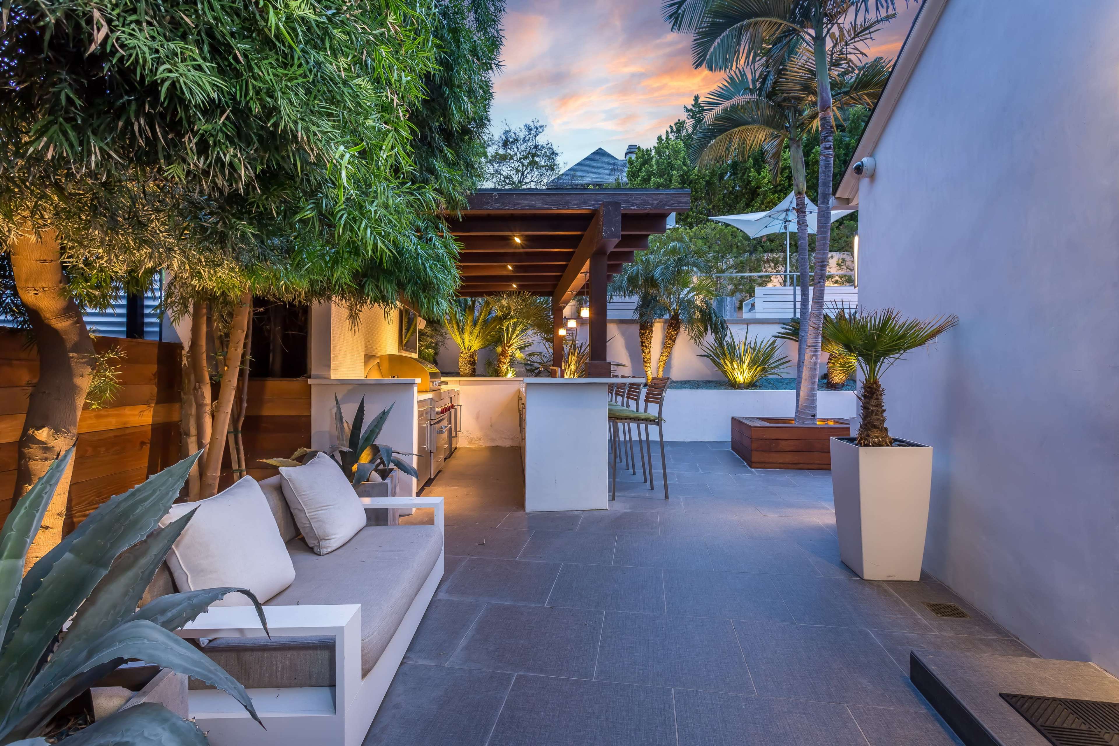 The image depicts a modern outdoor patio area featuring a bar, seating, and surrounding greenery under a colorful sky at dusk.