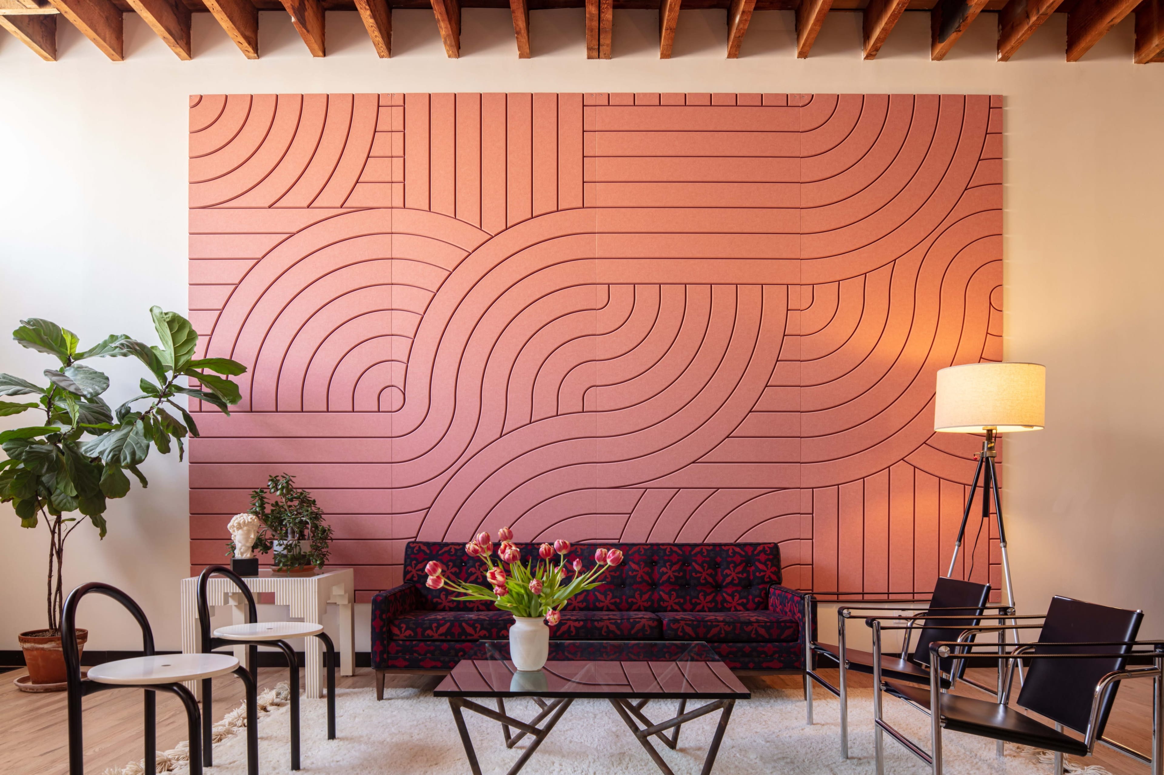 A modern living room with a pink, patterned accent wall, a dark sofa, several chairs, a glass coffee table, and a vase of flowers on display.