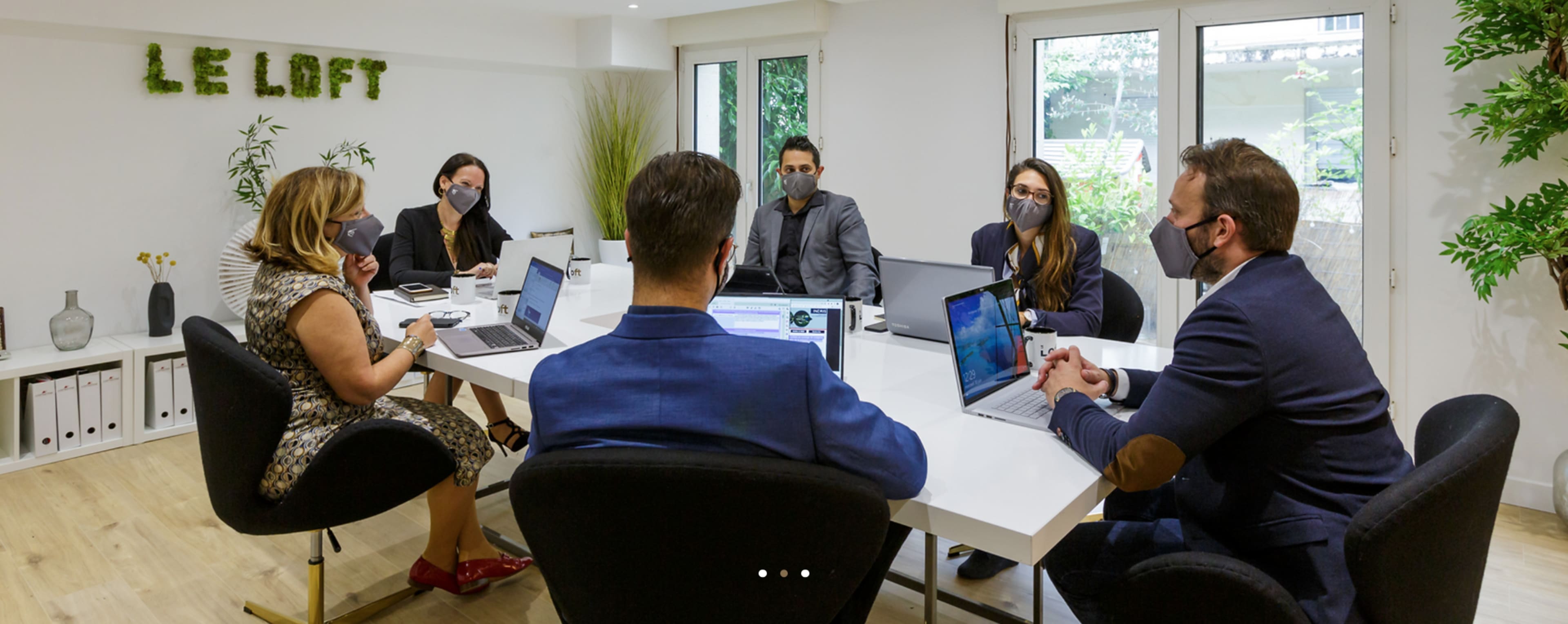 A group of six people wearing masks is seated around a conference table with laptops and documents in a well-lit meeting room.