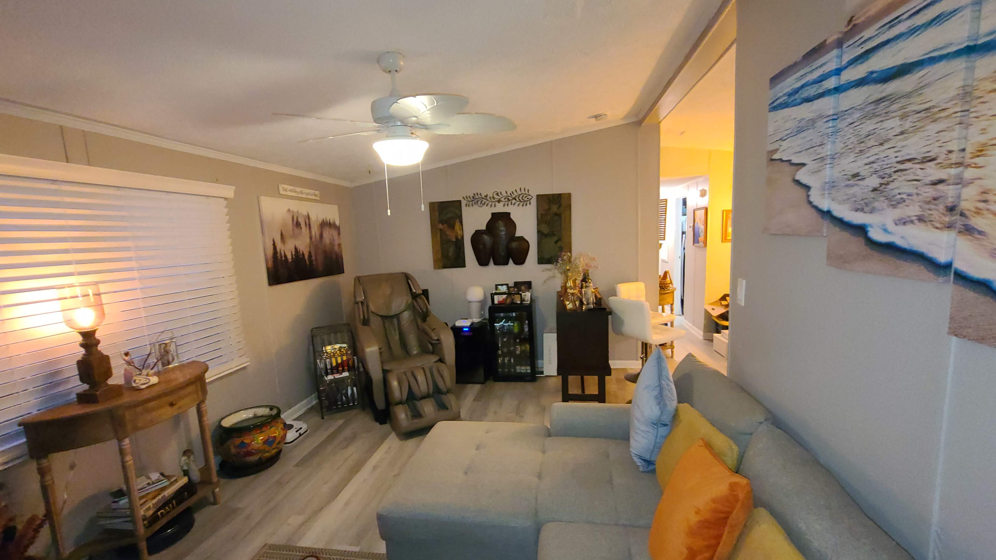 The image shows a cozy living room with a couch, a massage chair, and a small table, illuminated by a ceiling fan and soft lamps.